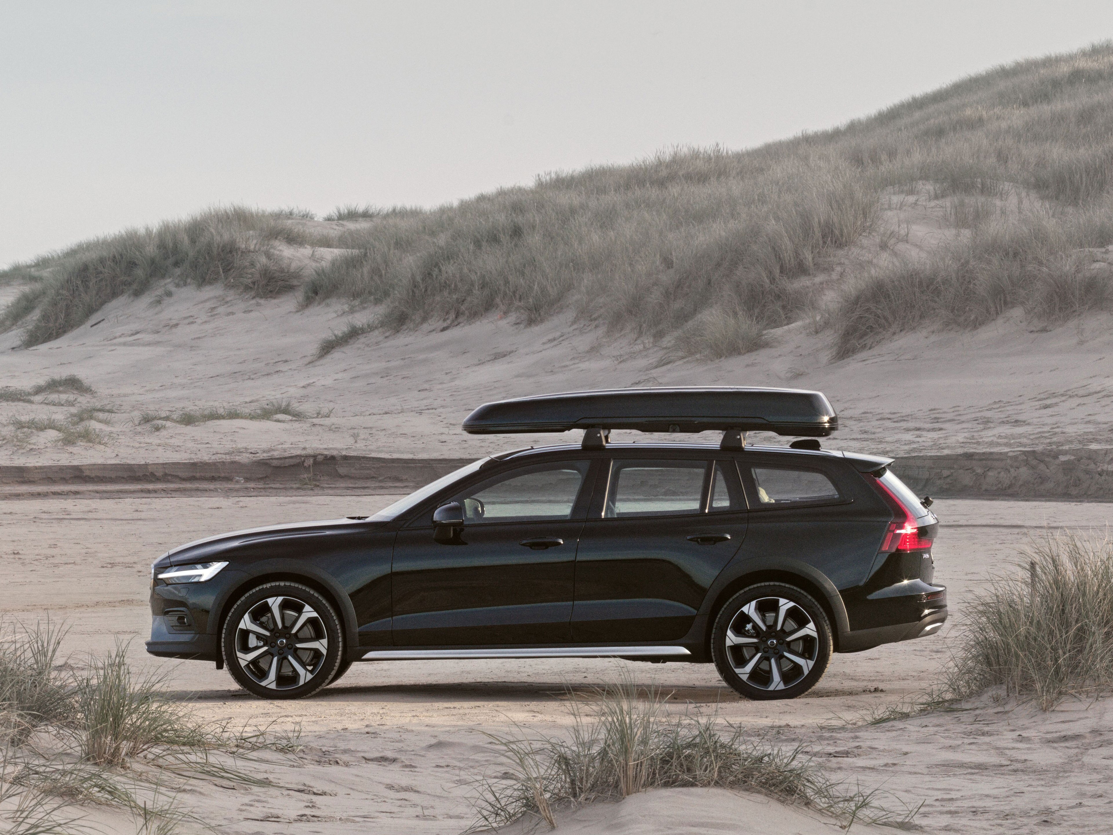 Ein schwarzer Volvo V60 Cross Country mit Dachbox steht auf einem Stück Sandstrand vor einer Düne.