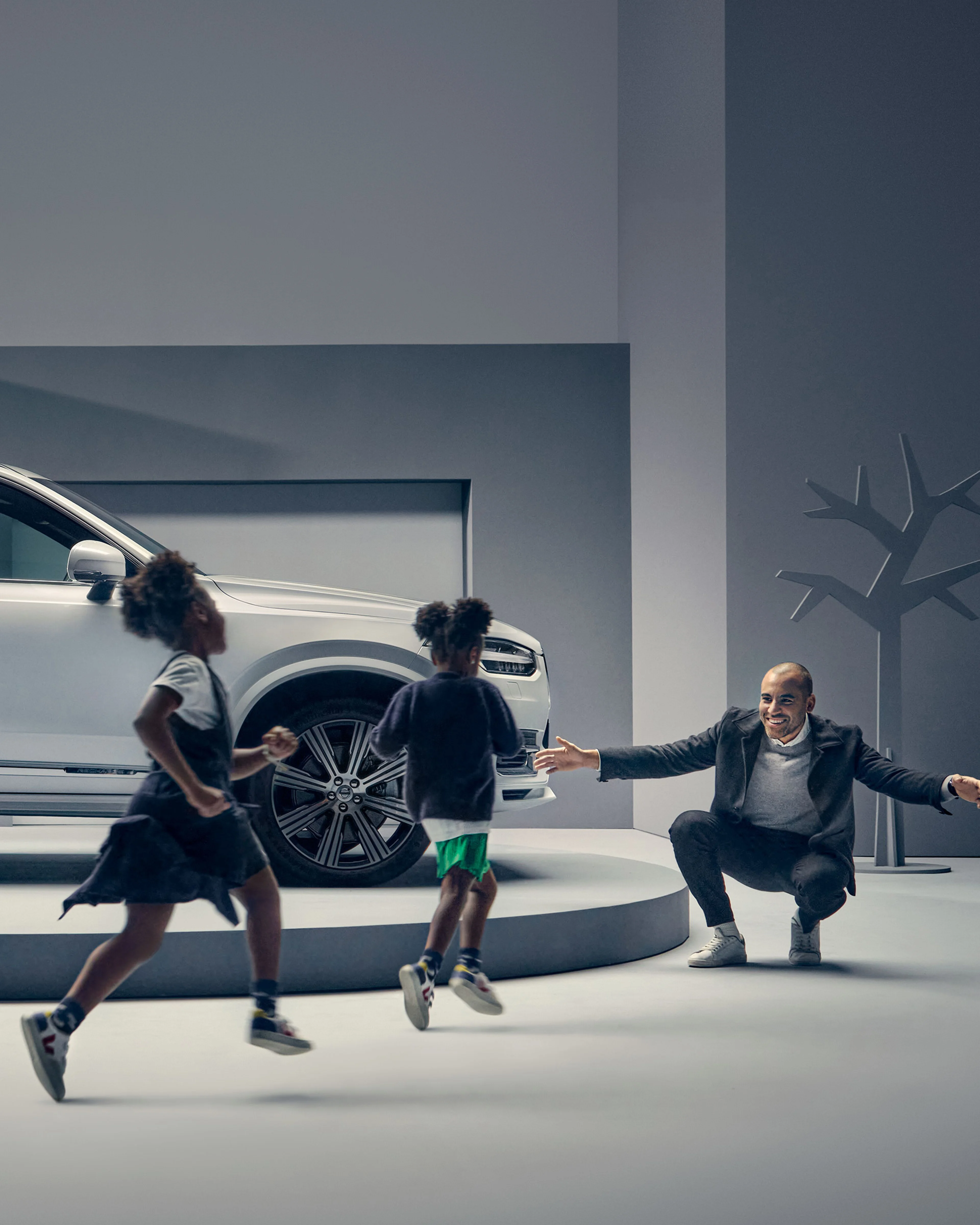 Un homme est assis, accroupi, les bras ouverts, à côté d'une Volvo XC90 tandis que deux enfants marchent vers lui.
