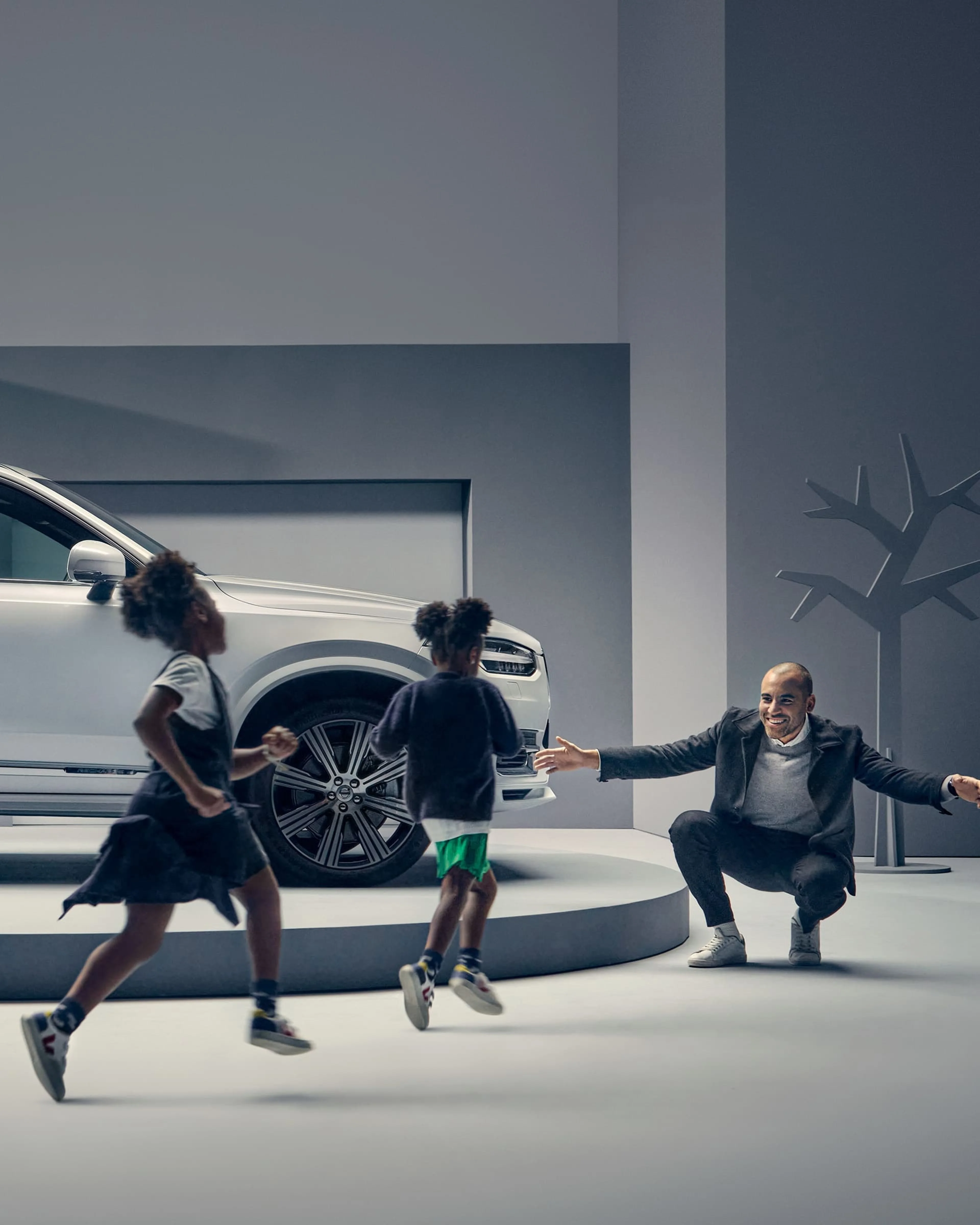 Un homme est assis, accroupi, les bras ouverts, à côté d'une Volvo XC90 tandis que deux enfants marchent vers lui.