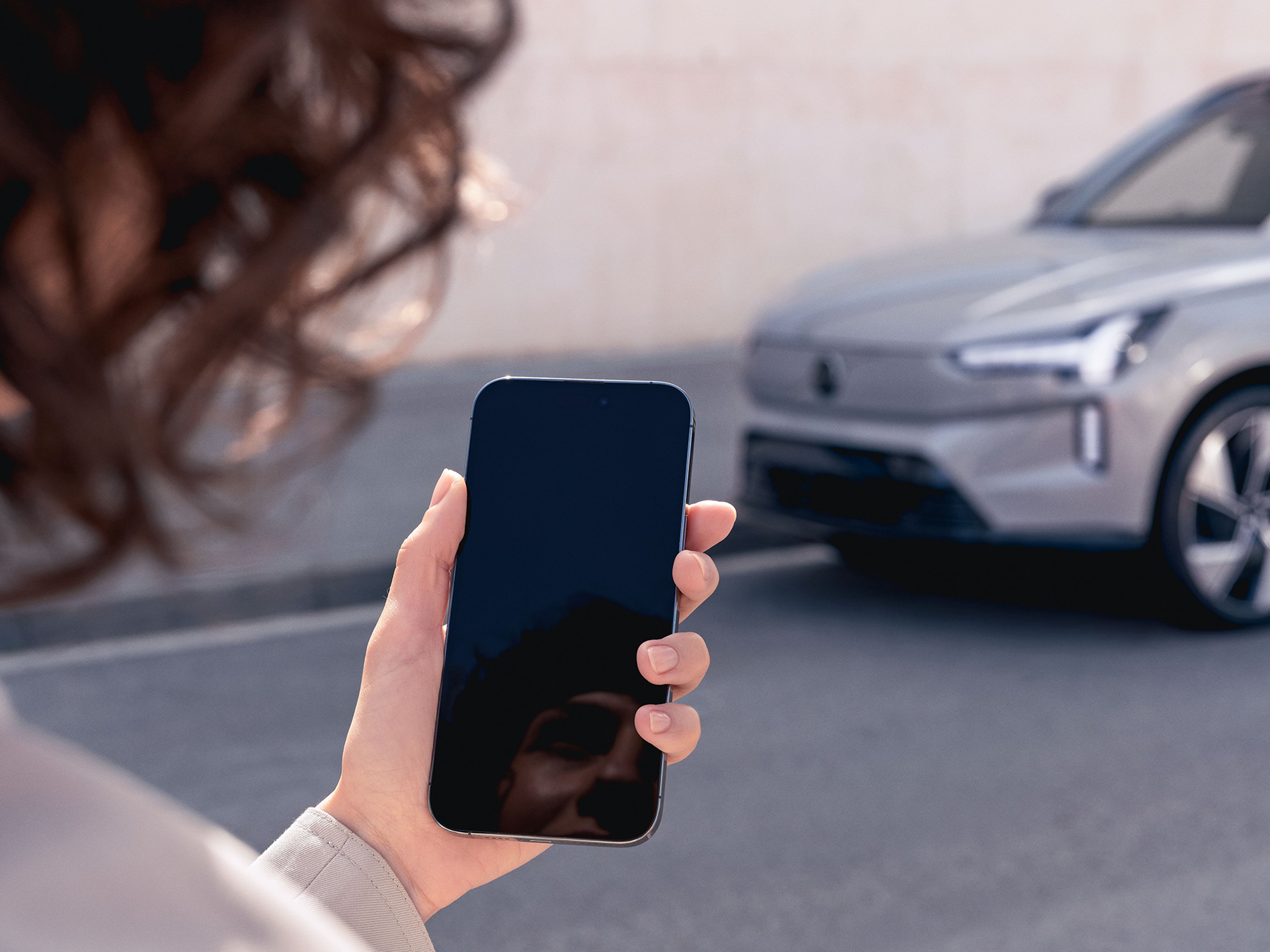 Kvinna som håller i en mobil med en grå Volvo i bakgrunden till höger.