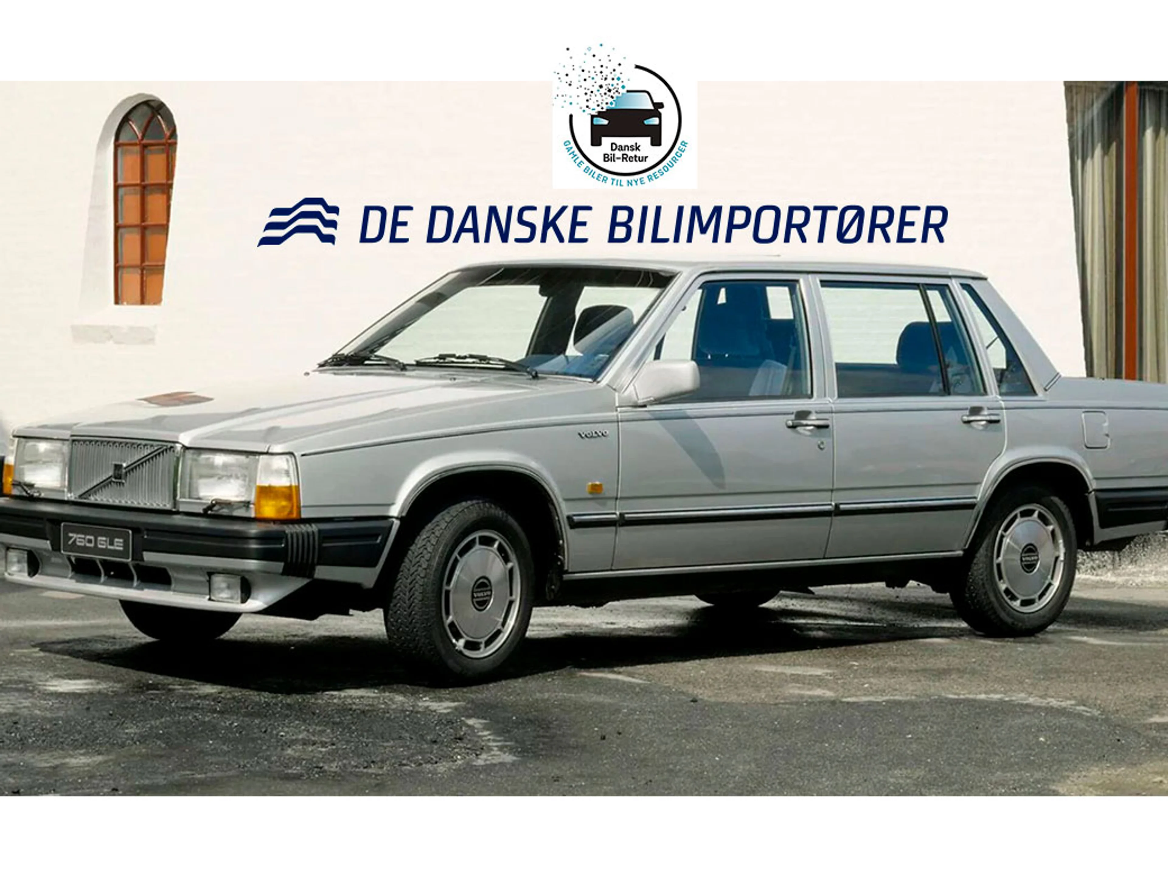 En sølvfarvet Volvo 760 GLE sedan holder parkeret foran en hvid mur med buede vinduer. Logoet og navnet "De Danske Bilimportører" er vist ovenover.