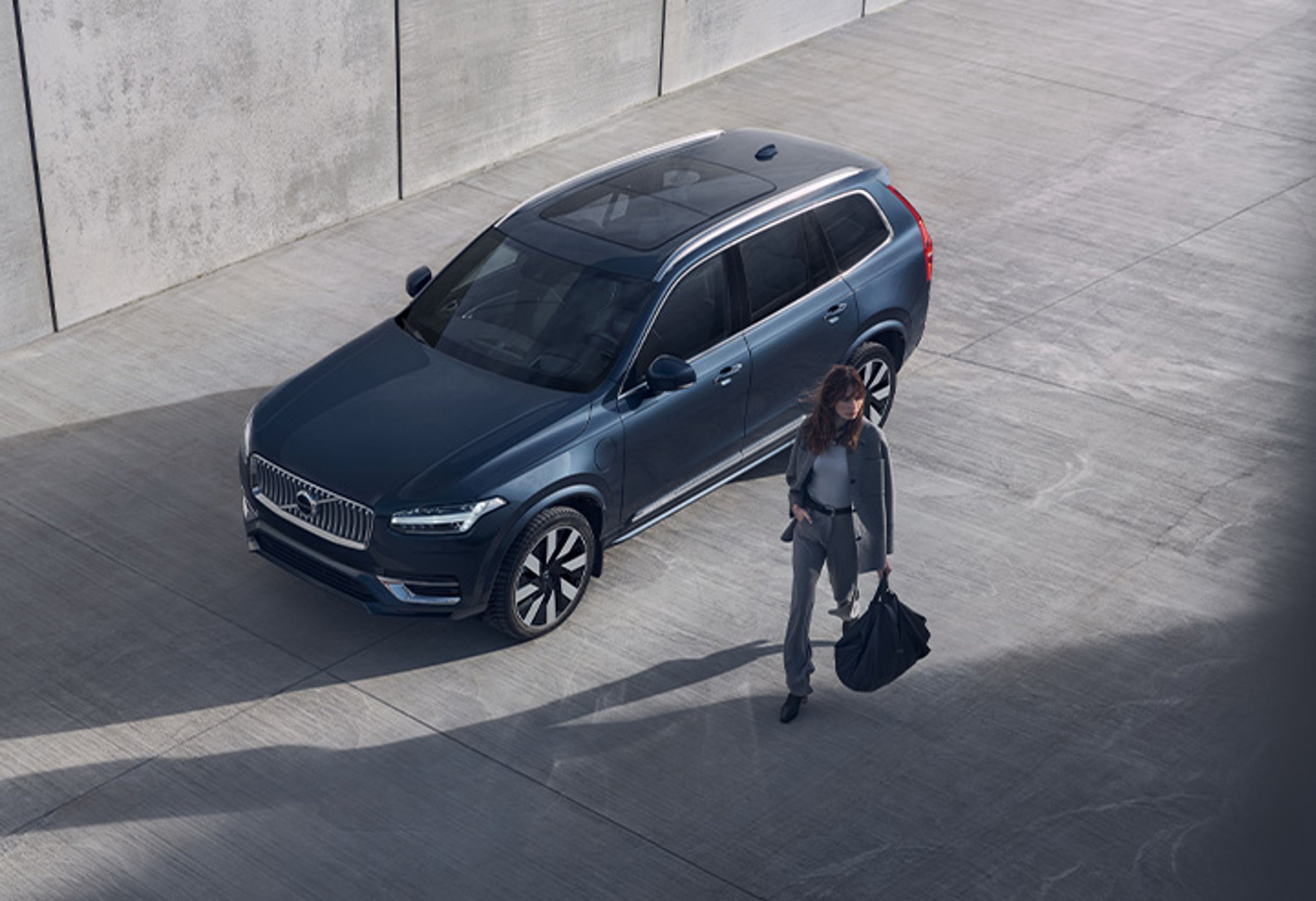 Eine Frau mit grauem Anzug und schwarzer Tasche in der Hand geht an einem dunkelblauen Volvo XC90 SUV vorbei.
