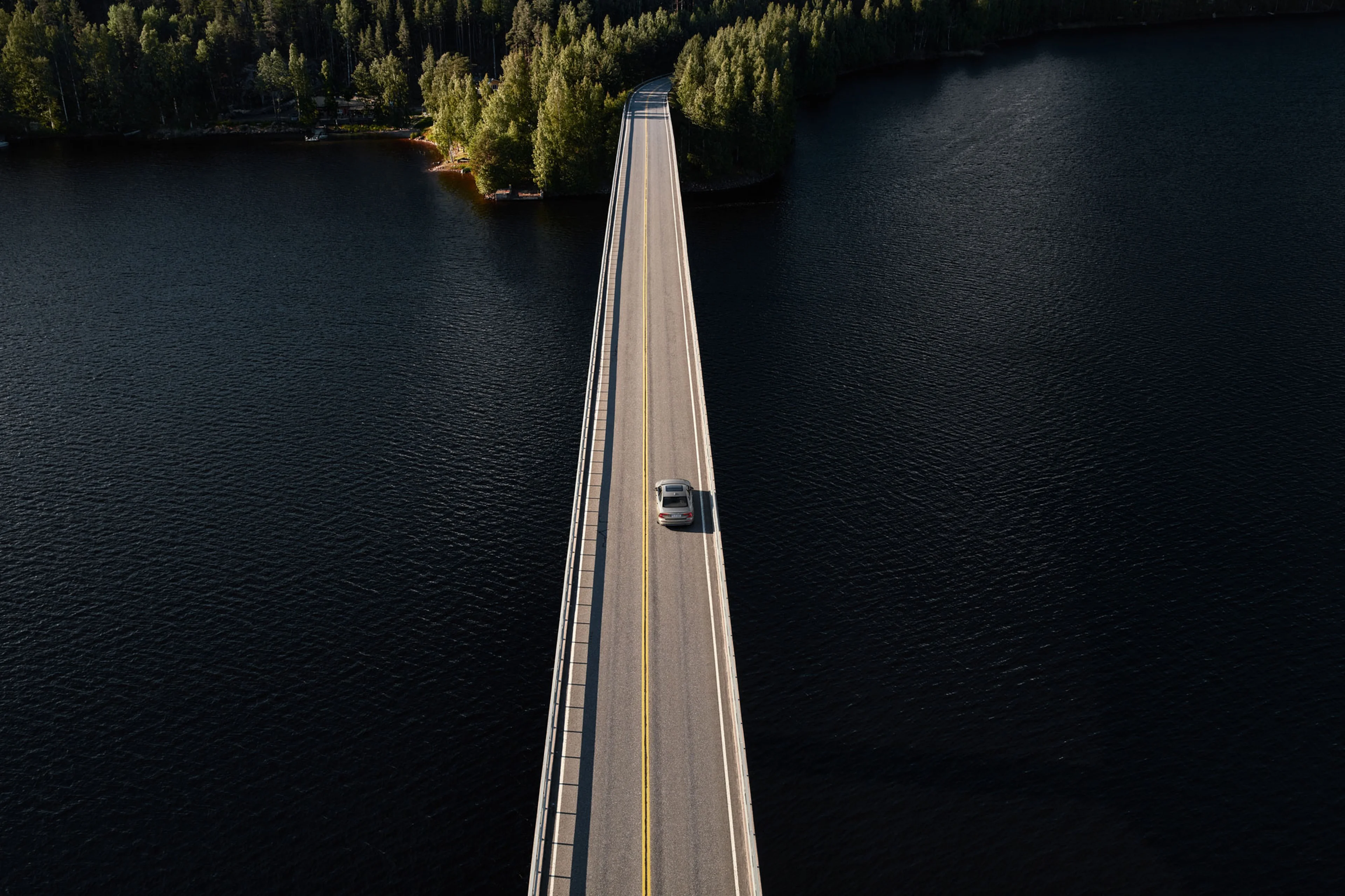 Luftfoto af en sølvfarvet Volvo, der kører over en lang, smal bro over mørkt vand, omgivet af frodige grønne skove. Scenen udstråler ro og afsondrethed.