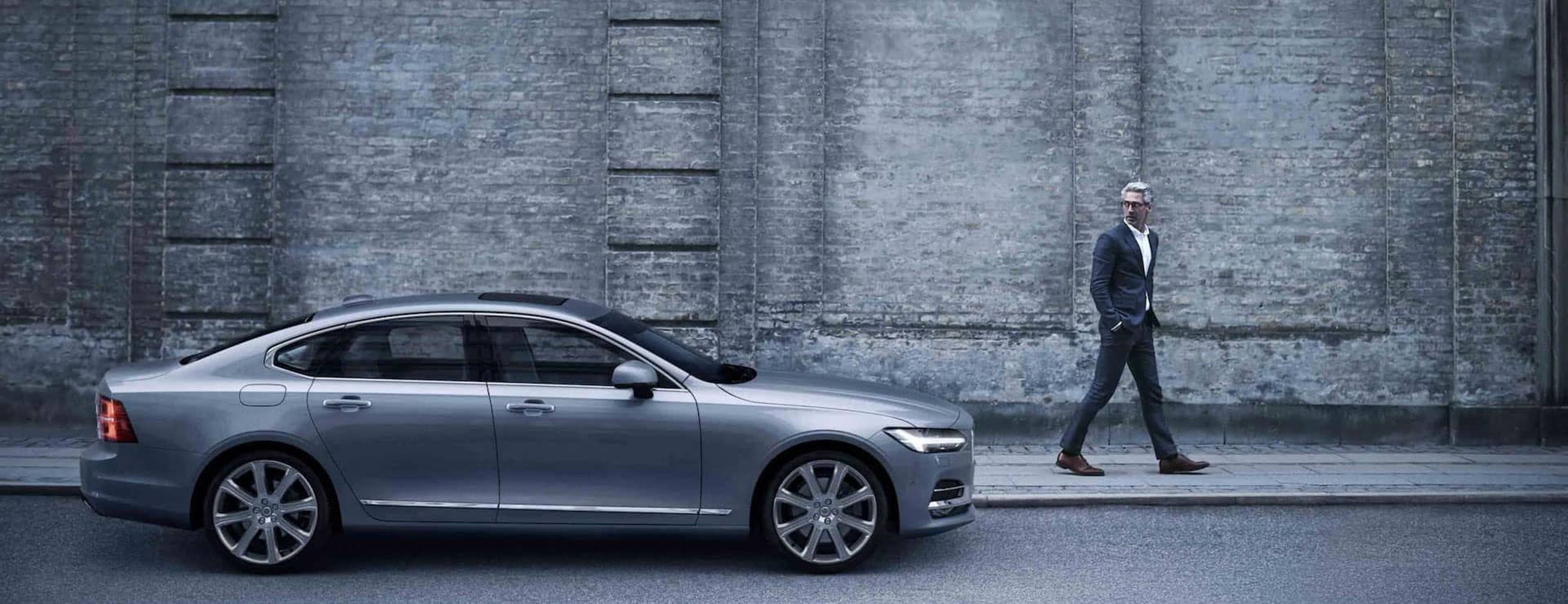 A grey Volvo S90 parked by the sidewalk in front of an old brick wall, with a man in a suit walking past it.