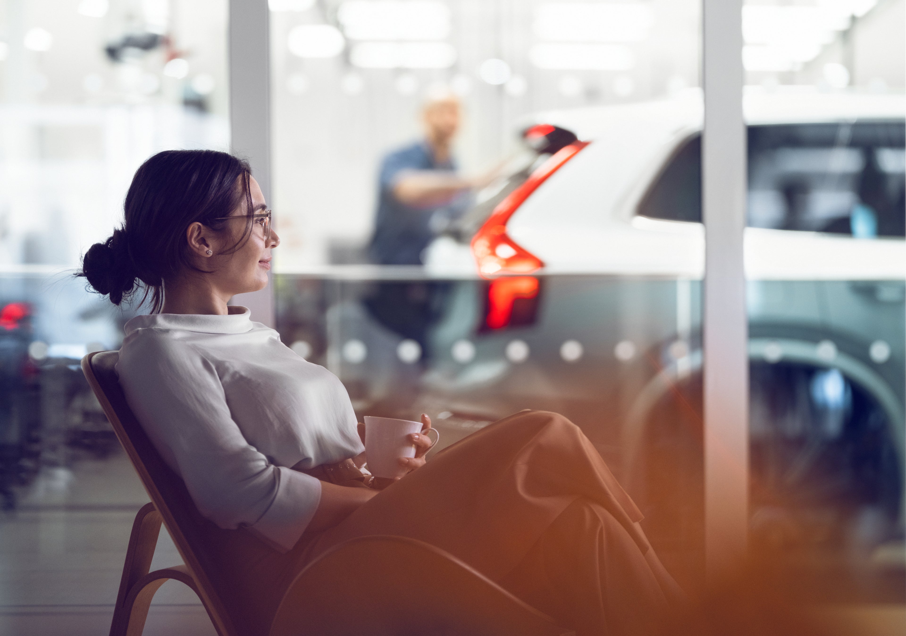 Жена в профил, удобно настанена в стол с чаша кафе в ръка, на заден план Volvo XC90 преминава сервизна проверка от техник.