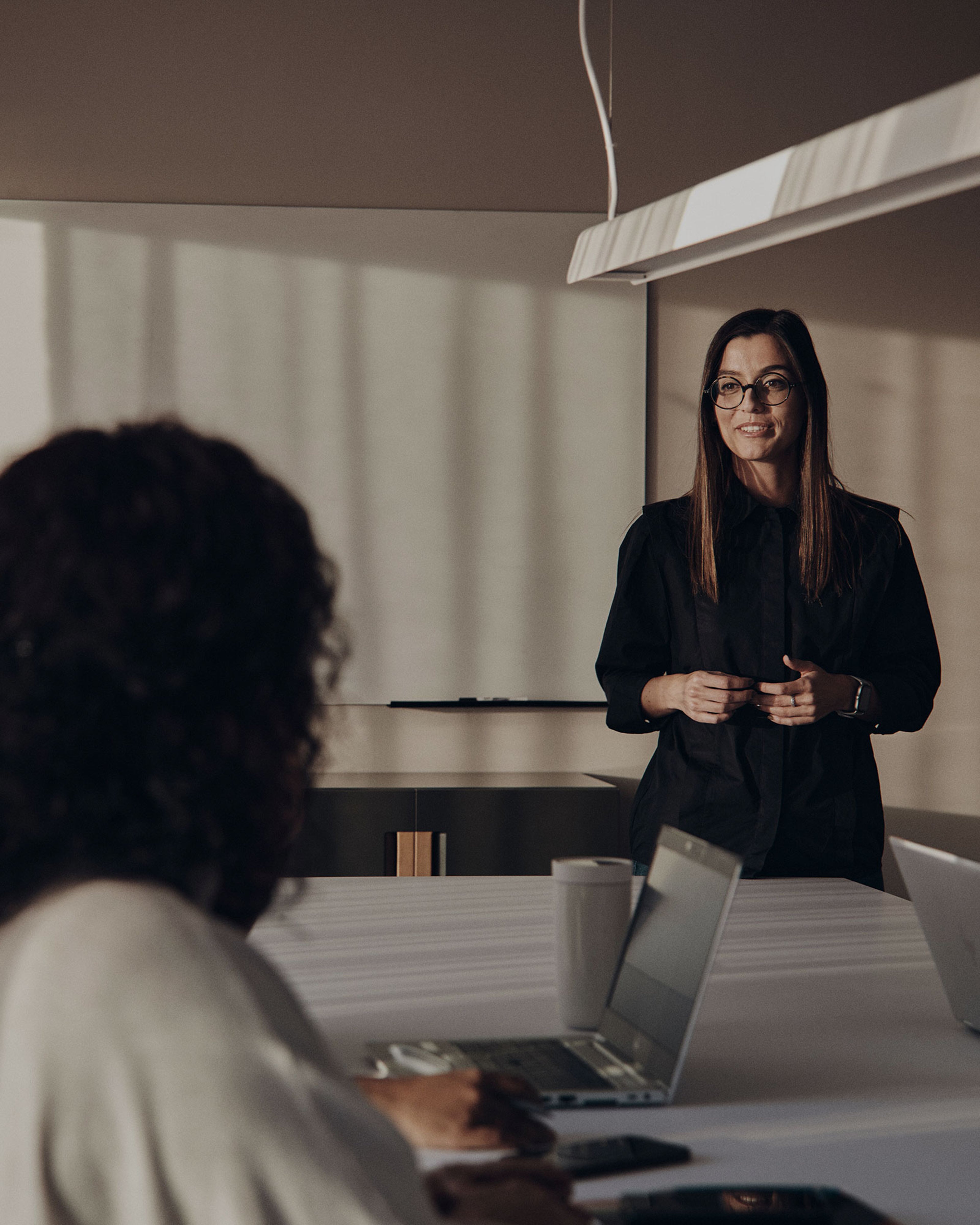 Een vrouw met een bril en een zwart overhemd aan een tafel en praat tegen een andere persoon in een lichte kantoorruimte.