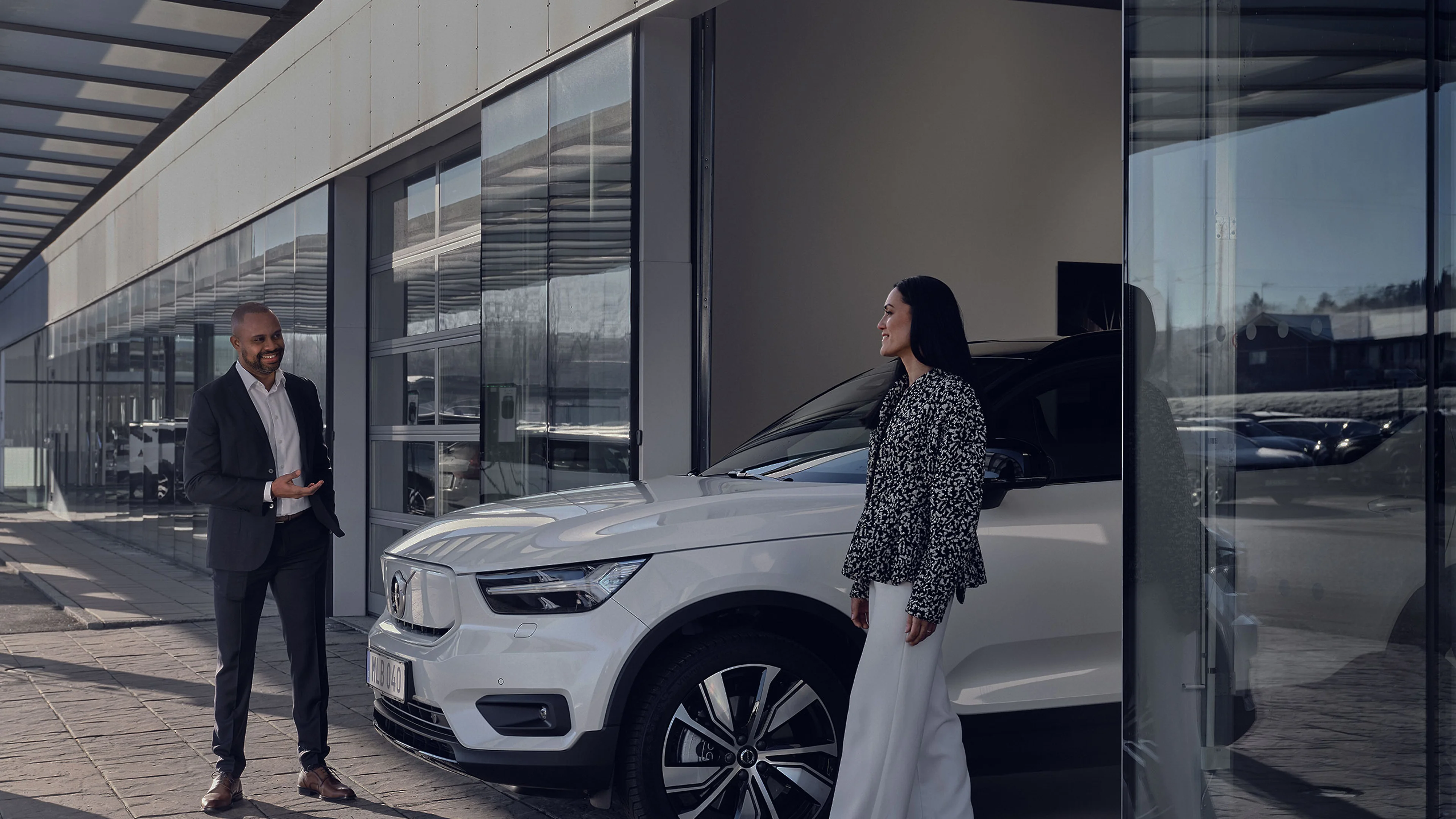 En mand i jakkesæt smiler, mens han taler med en kvinde i en mønstret bluse ved siden af en hvid Volvo-bil. Omgivelserne er et moderne Volvo-bilhus med glasvægge.