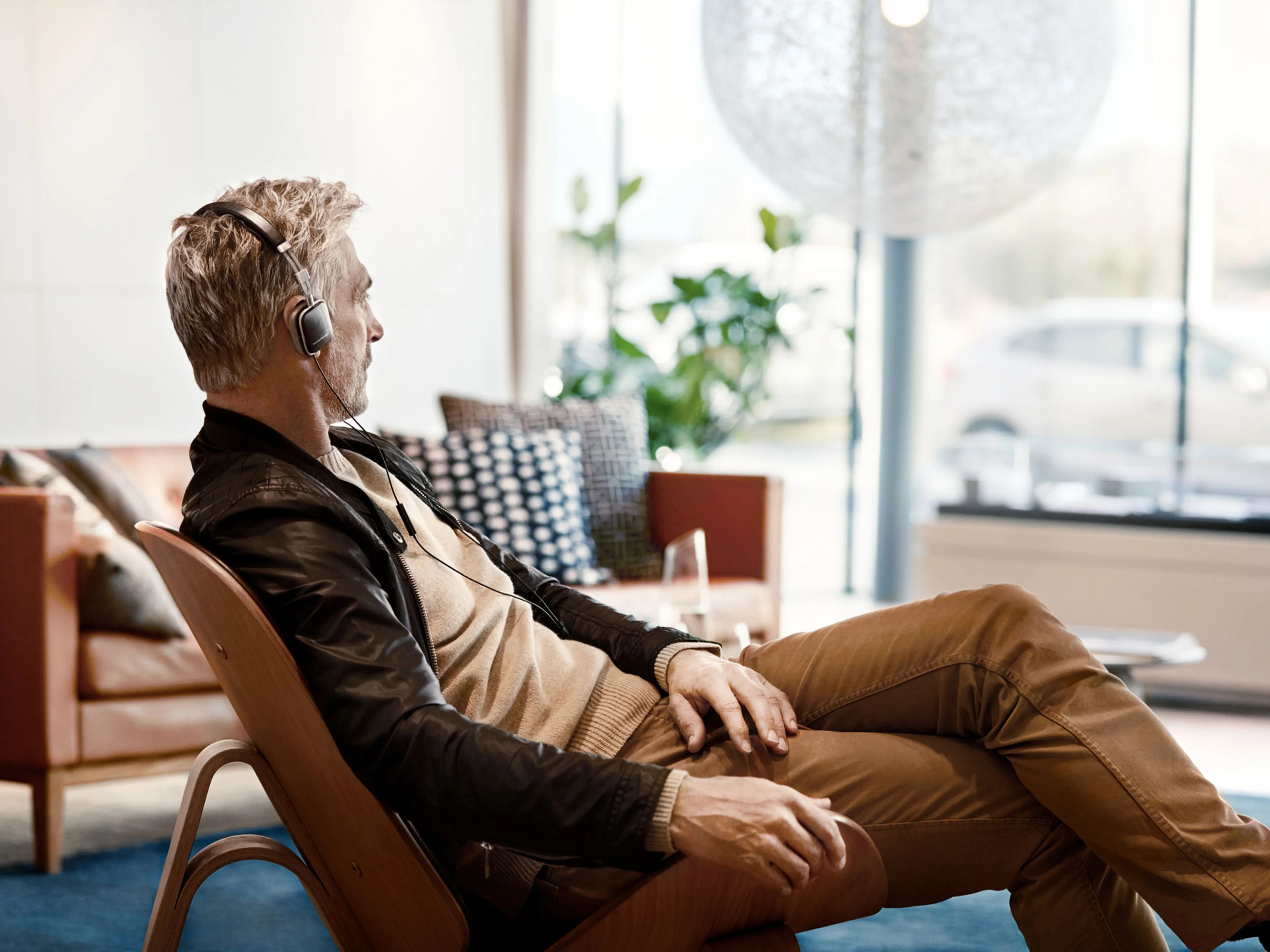 En mand med gråt hår sidder i et moderne, stilfuldt rum hos en Volvo forhandler iført hovedtelefoner. Han ser afslappet ud og kigger ud gennem store vinduer.