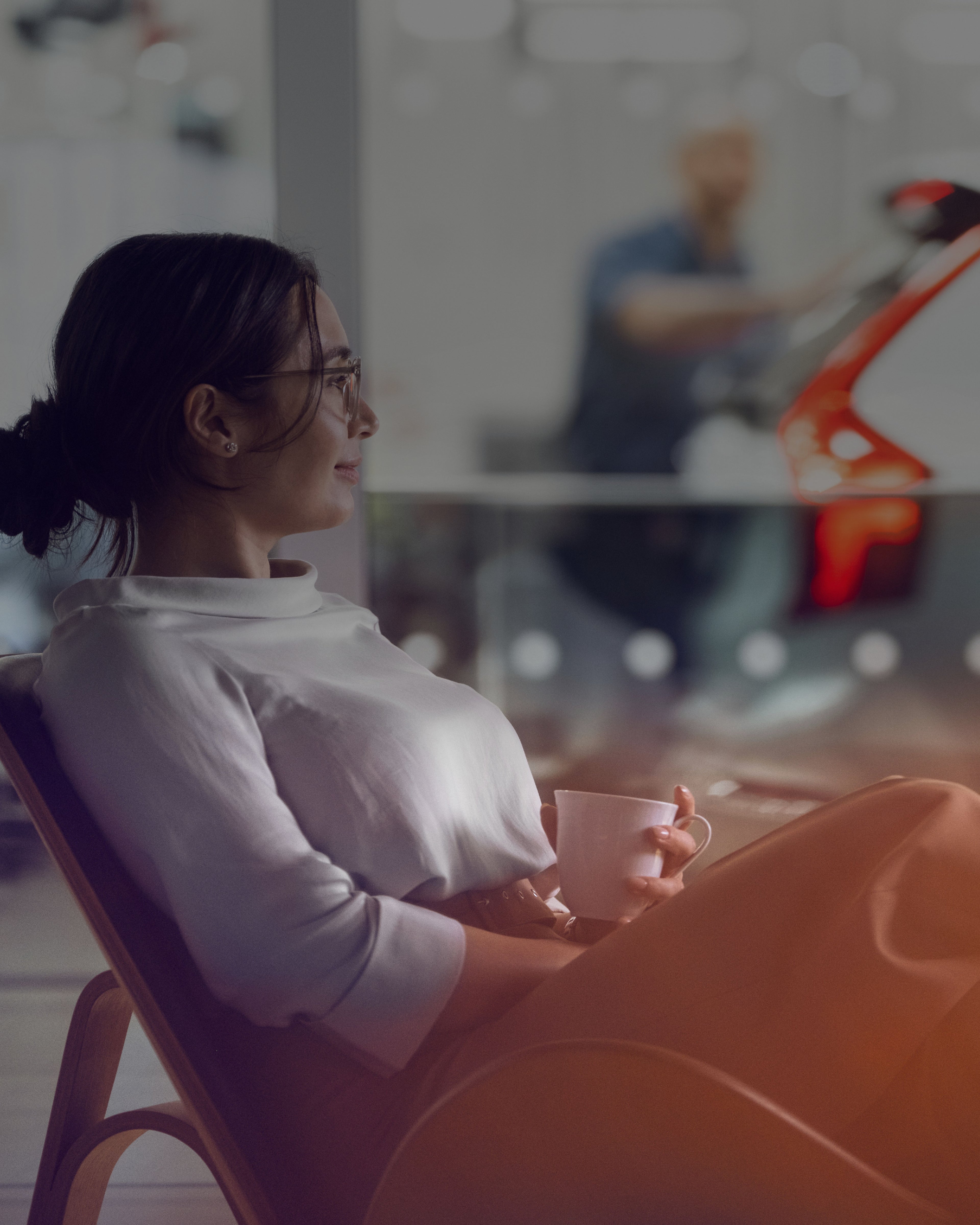 Eine Frau mit einer Tasse in der Hand sitzt in einer Werkstatt. Im Hintergrund ist ein Servicetechniker zu sehen.