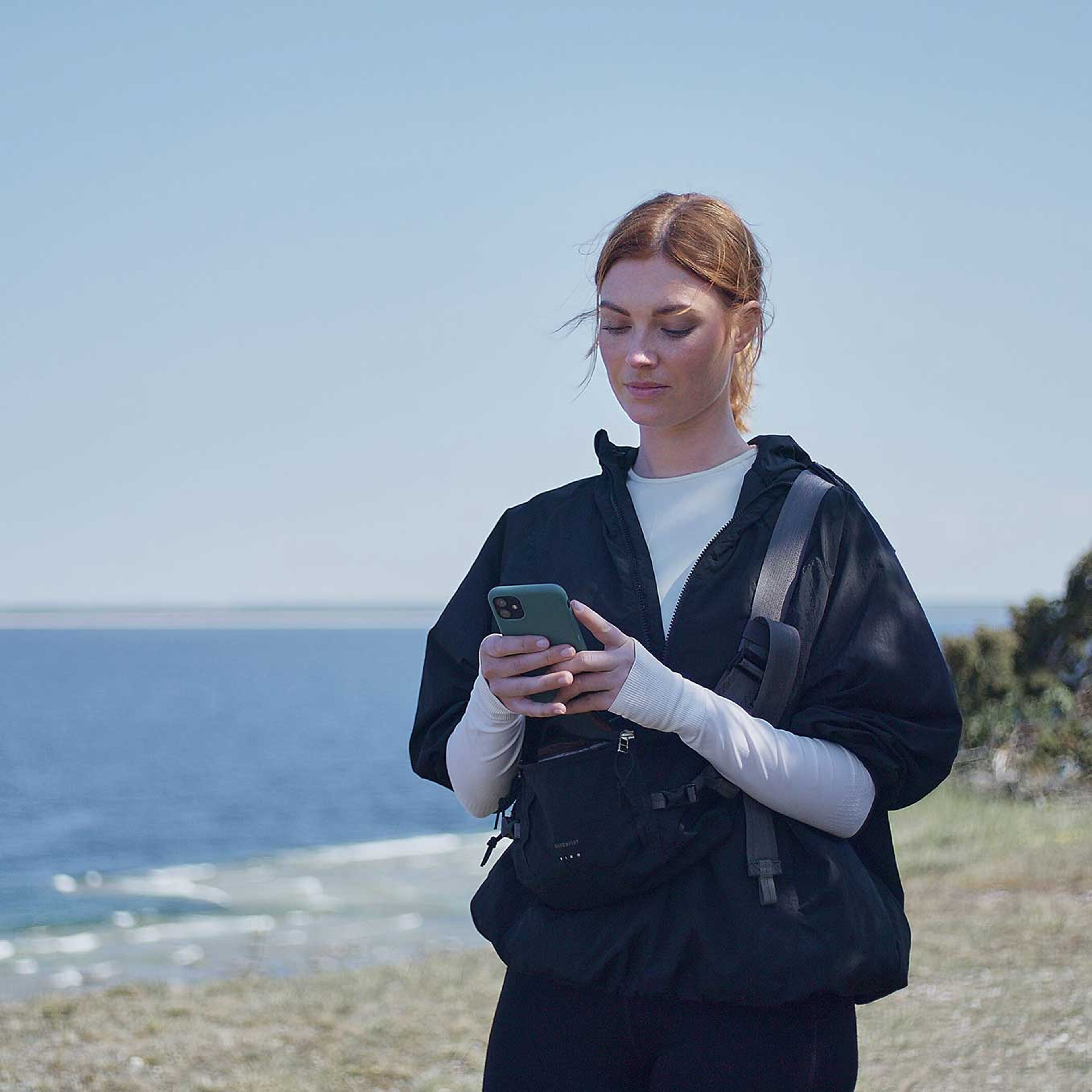 En kvinde ses lidt fra siden forfra mens hun kigger ned og skriver på sin smartphone. Hun er udenfor i et solrigt landskab med vand i baggrunden.