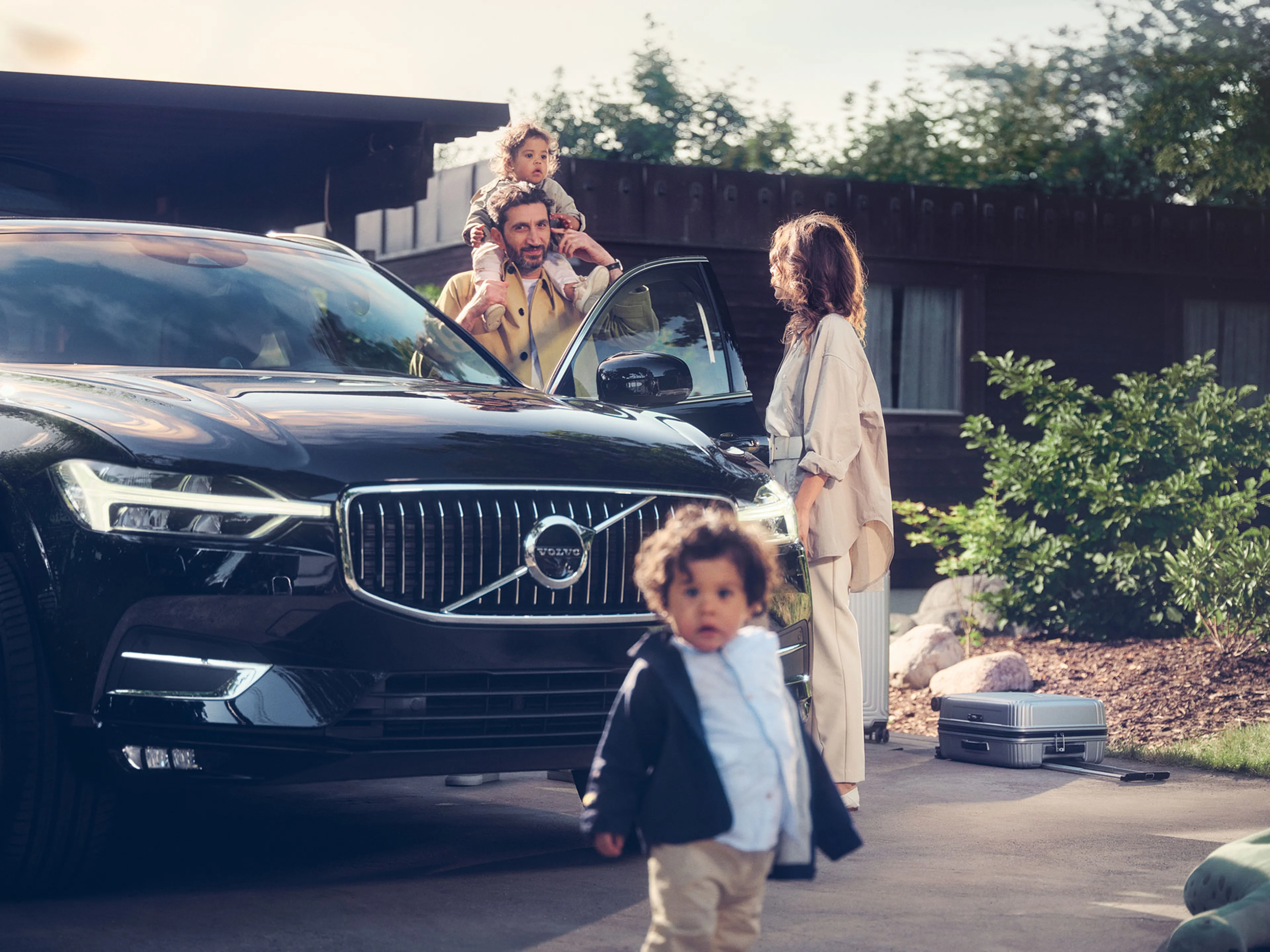 Een zwarte Volvo geparkeerd voor een huis, met een gezin: een vader met een kind op zijn schouders, een moeder die uit de auto stapt, en een kind dat naar de camera loopt. Er staat bagage naast de auto.
