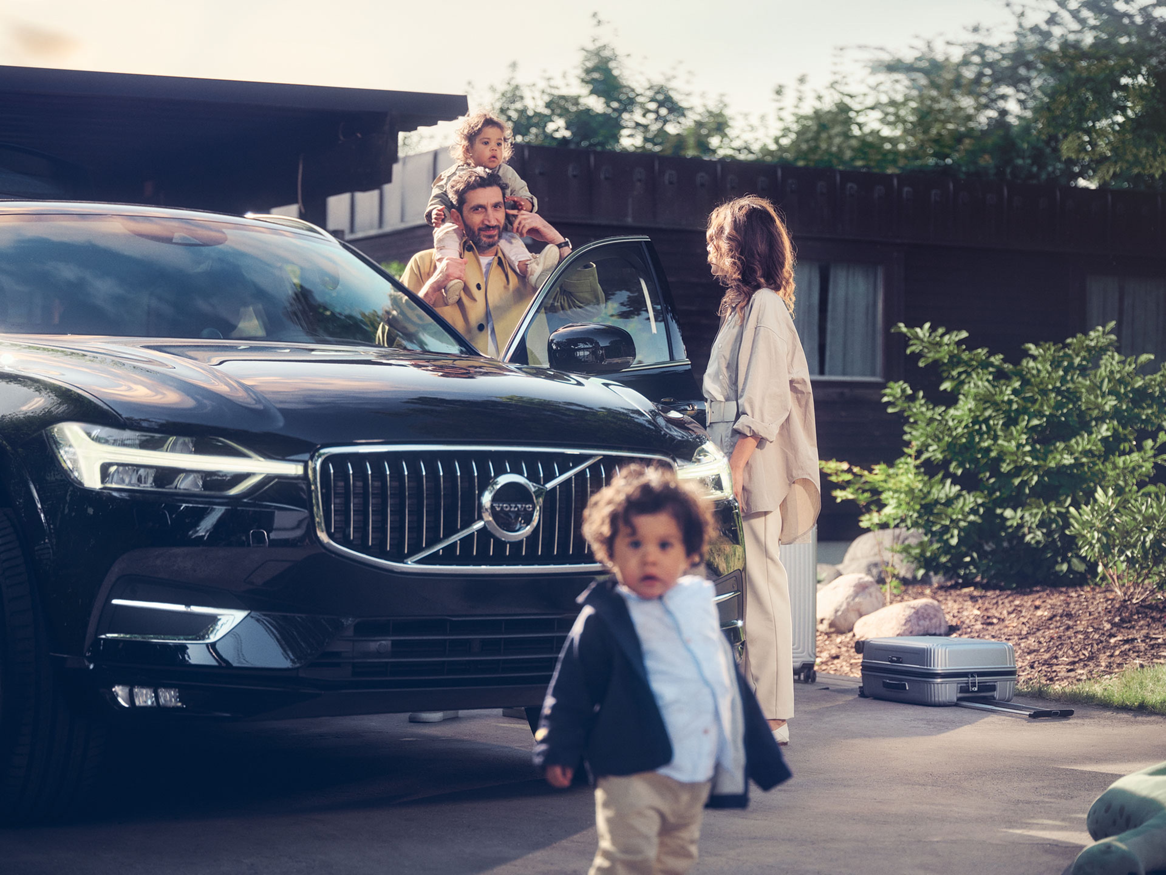 Une Volvo noire garée devant une maison, avec une famille : un père avec un enfant sur ses épaules, une mère sortant de la voiture, et un enfant marchant vers la caméra. Des bagages sont à côté de la voiture.