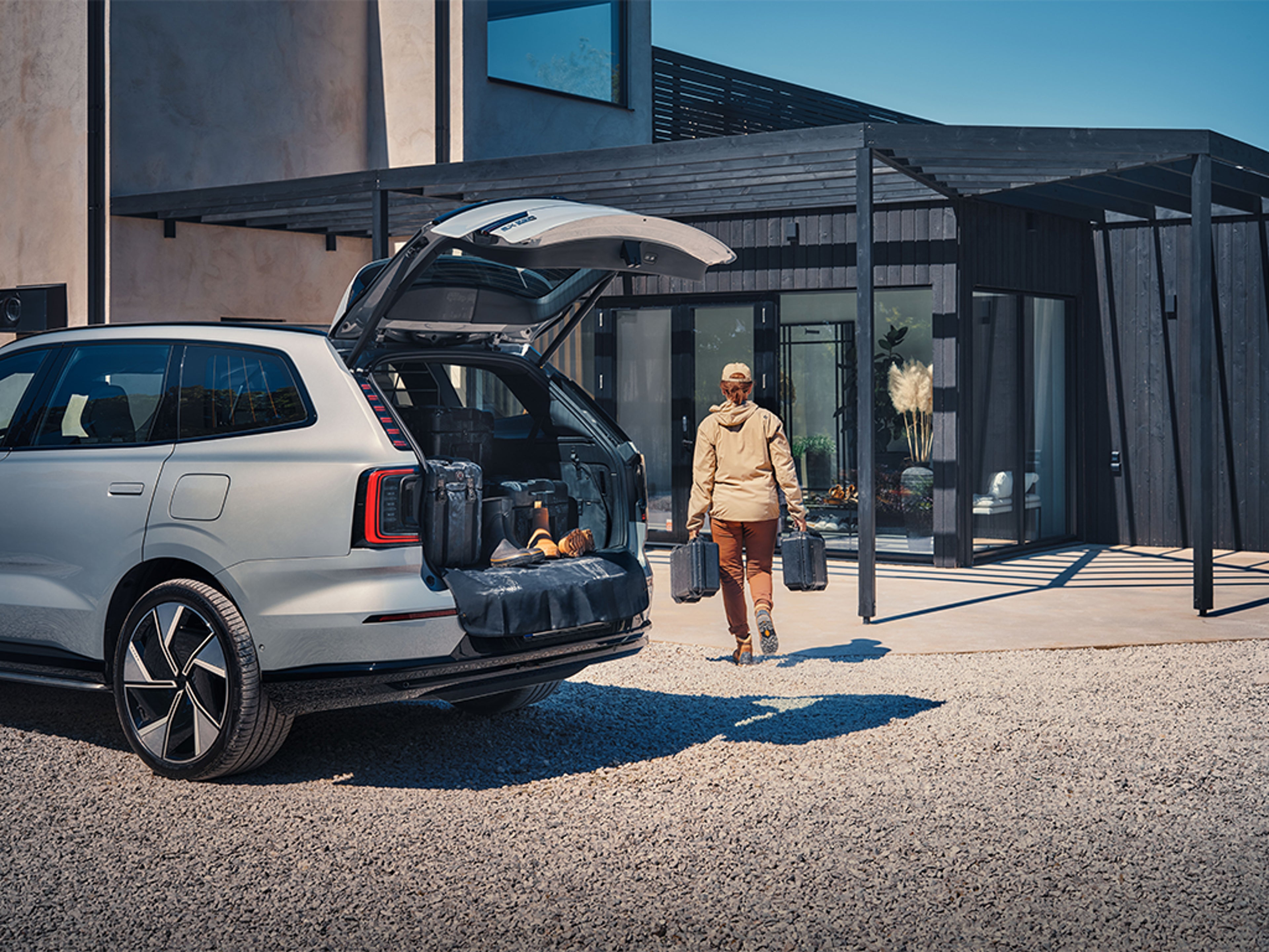 Volvo EX90 avec coffre ouvert, bagages à l’arrière, personne portant des valises devant une maison moderne