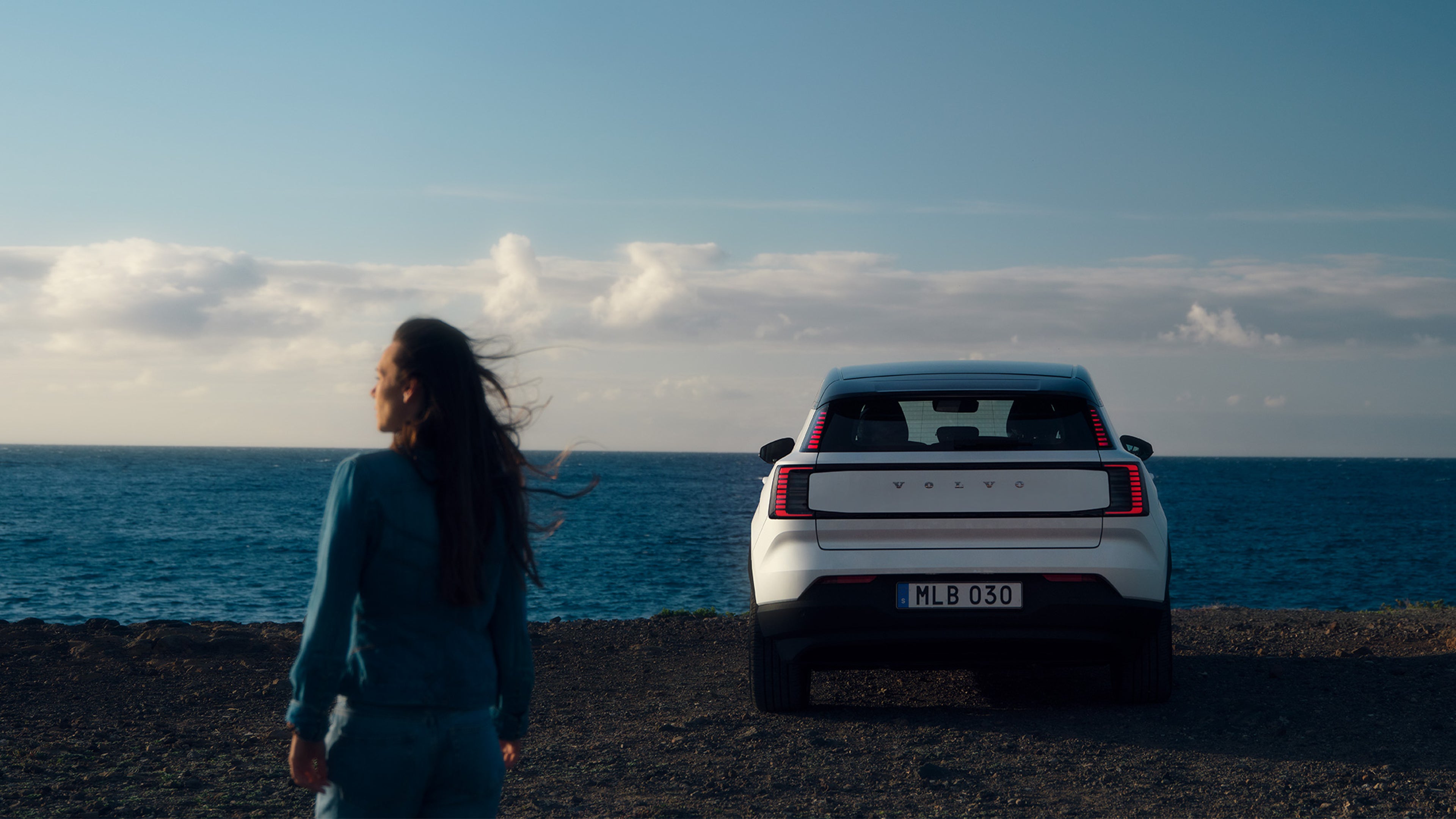 Merenrantaan pysäköidyn Crystal white -värisen Volvo EX30 täyssähköisen katumaasturin vieressä seisoo ihminen, jonka pitkä tumma tukka hulmuaa tuulessa. Taustalla meri ja ilta-aurinko.