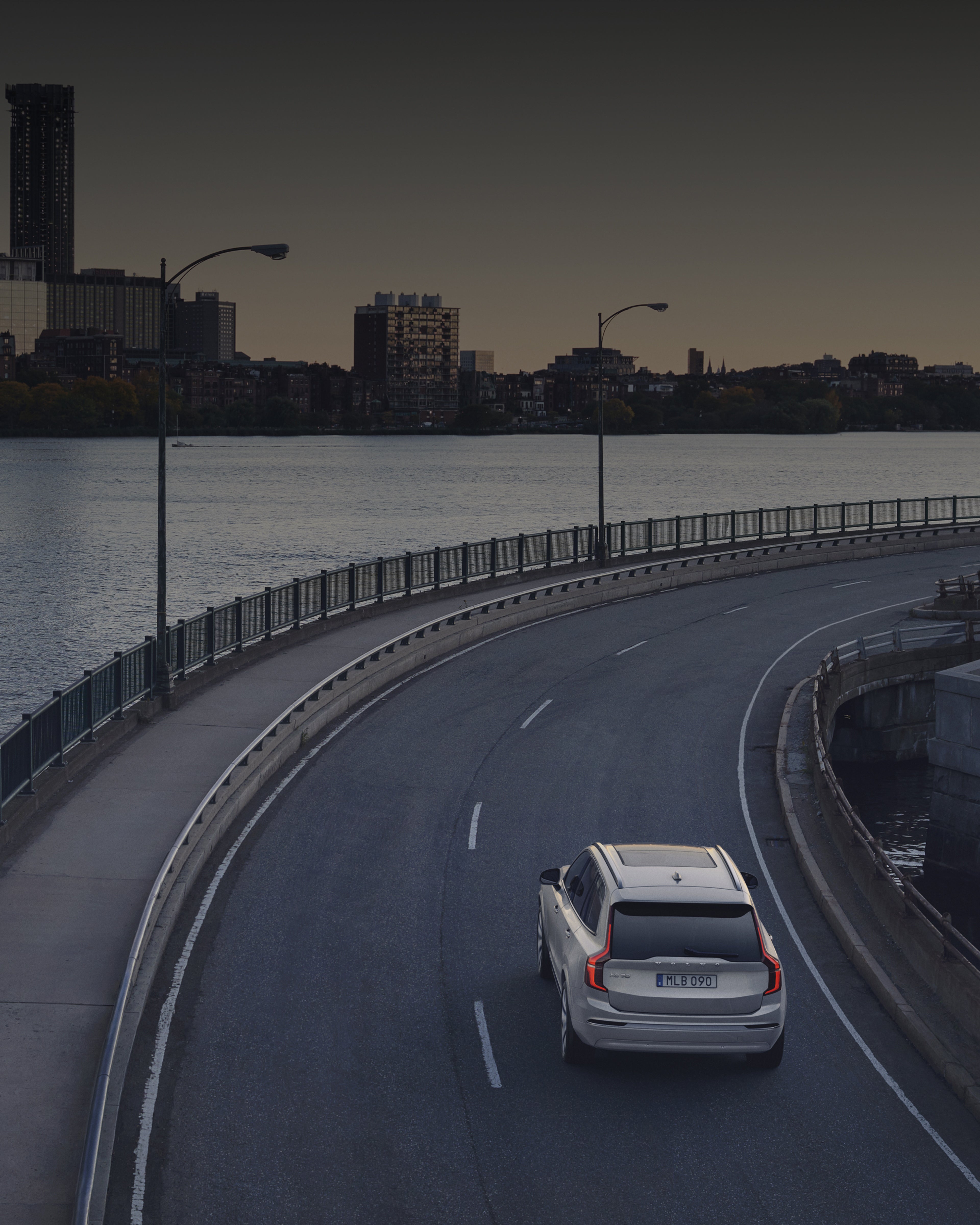 Un Volvo XC90 sobre una carretera con un río al lado y una ciudad de fondo.