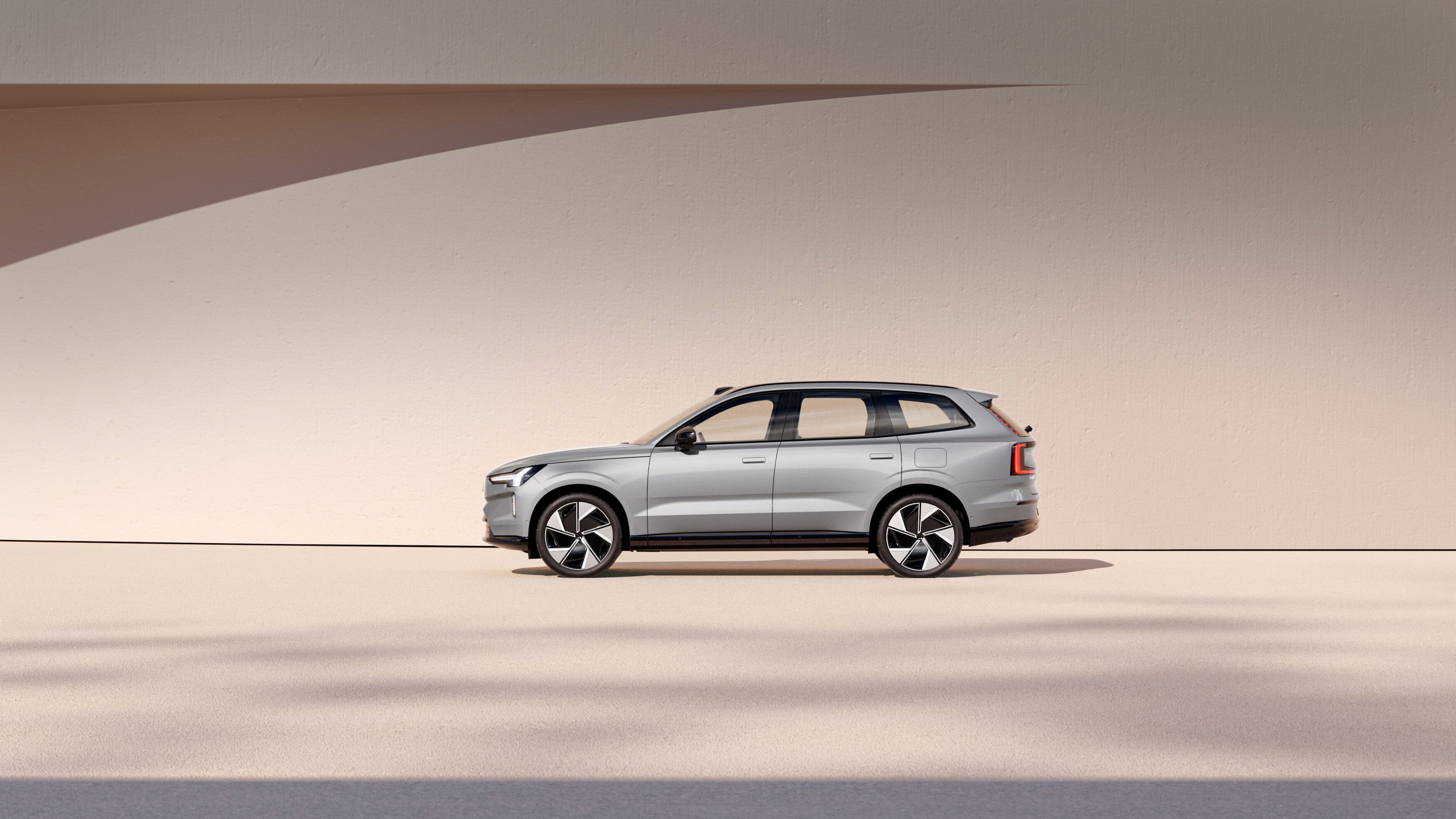 A side profile of the fully electric Volvo EX90 in Vapour Grey. Parked against a beige wall backdrop.