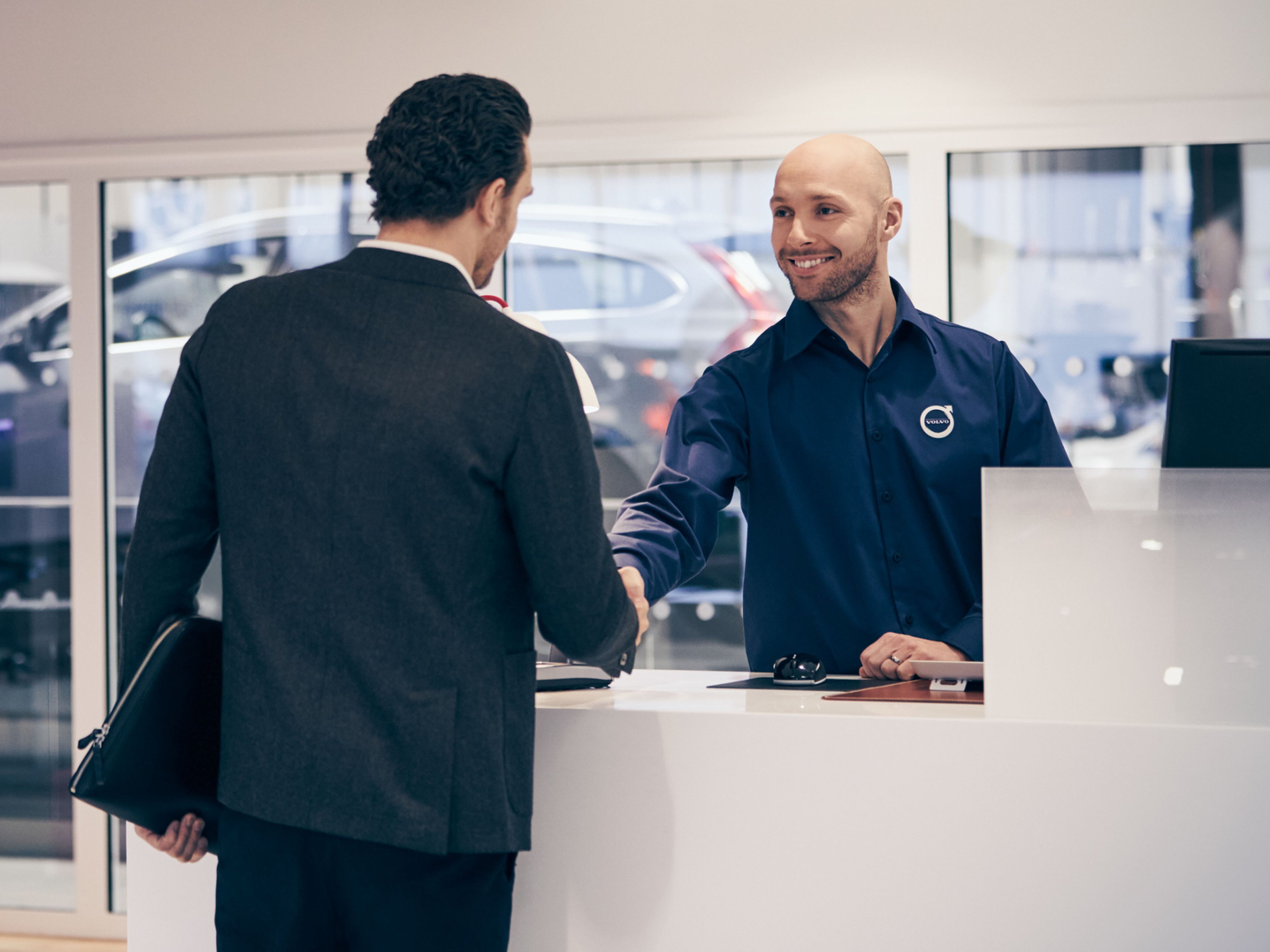 Ein männlicher Volvo Personal Service Techniker schüttelt einem männlichen Kunden die Hand. Im Hintergrund sieht man durch Glasscheiben eine Werkstatt mit einem Volvo XC90.