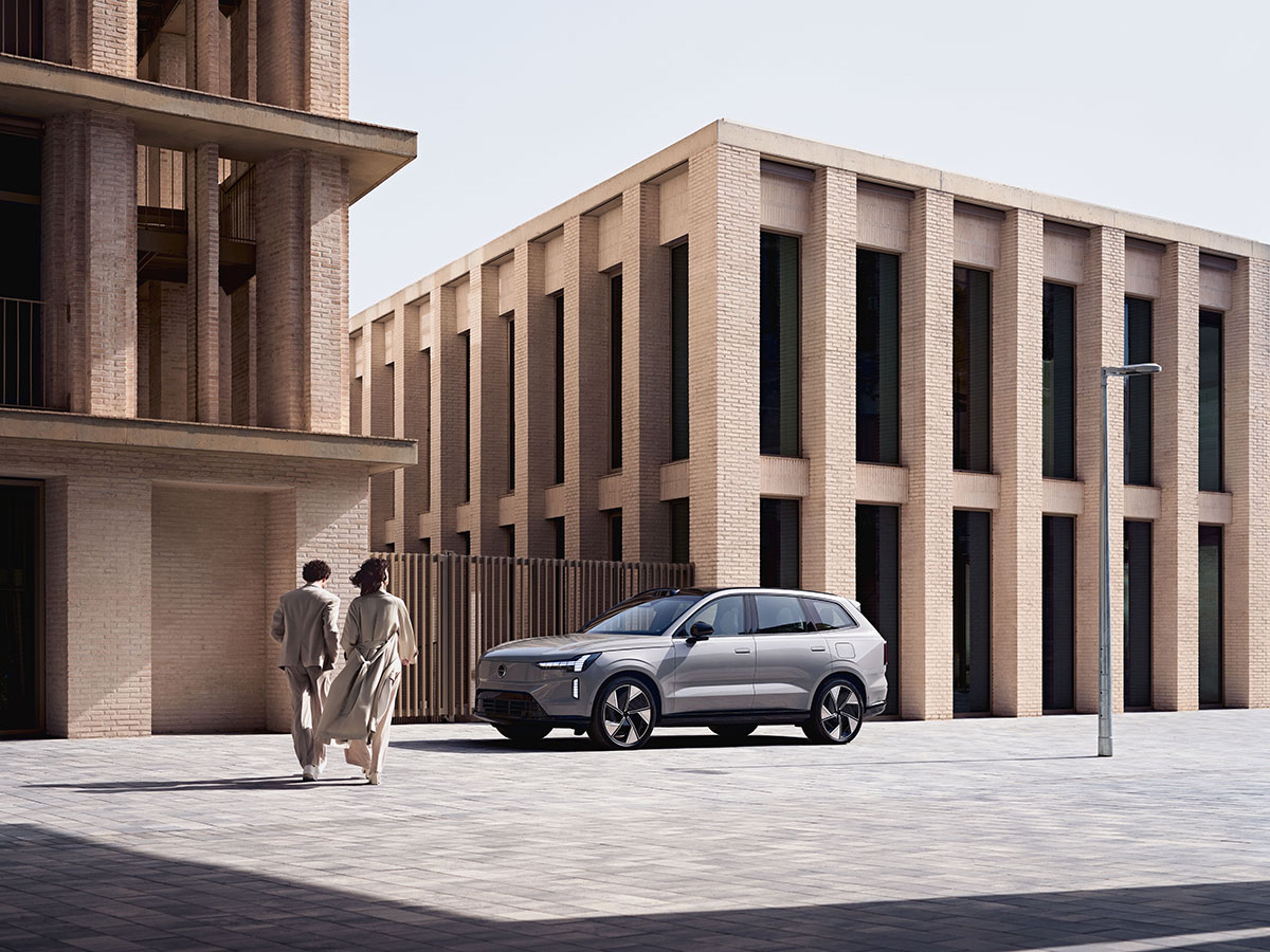 En sølvfarvet Volvo XC60 SUV er parkeret nær moderne bygninger i beige mursten. To personer i beige tøj går væk, hvilket skaber en rolig, urban stemning. 4o