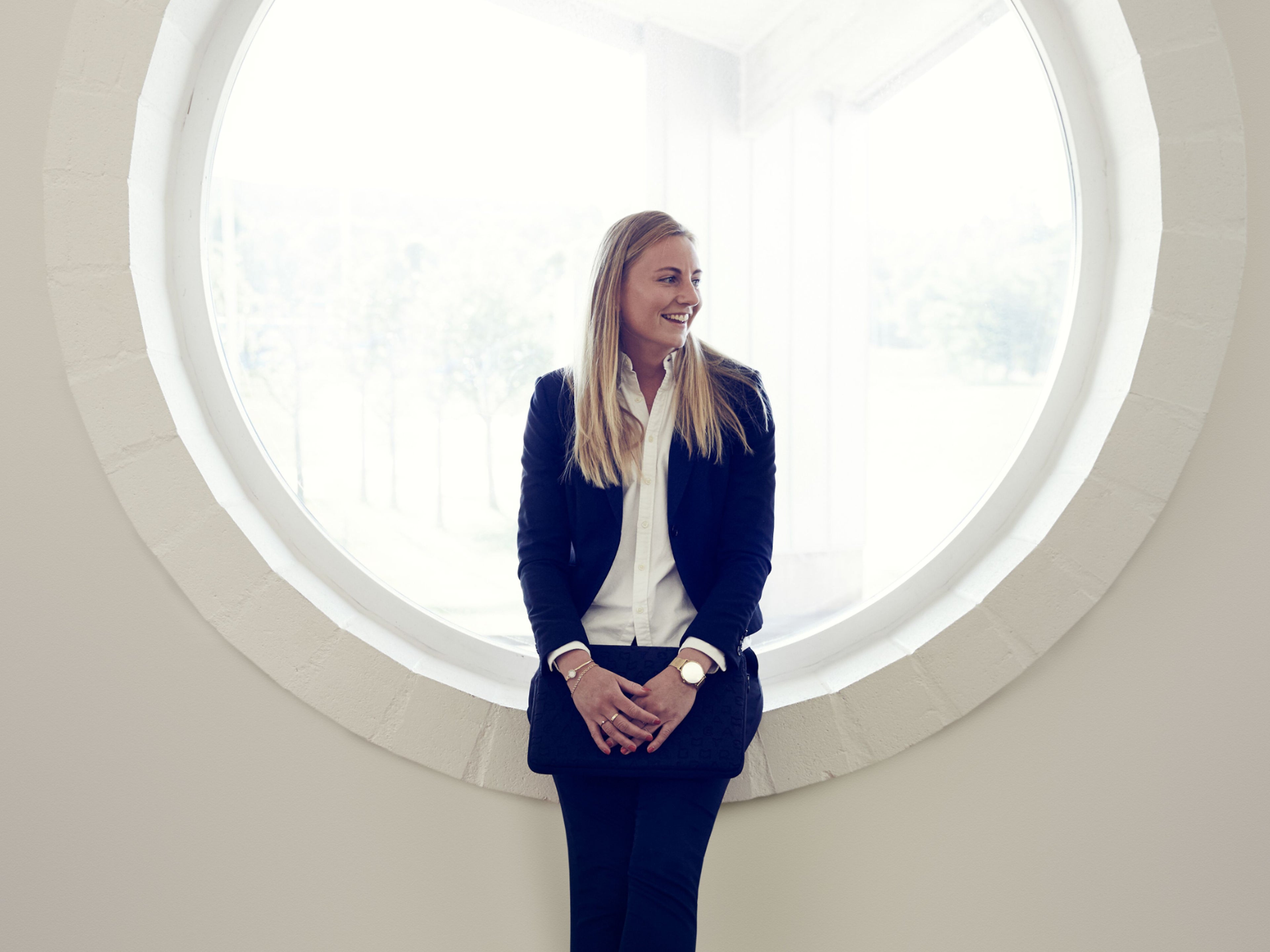 Eine Frau im Anzug steht vor einem runden Fenster und lächelt.