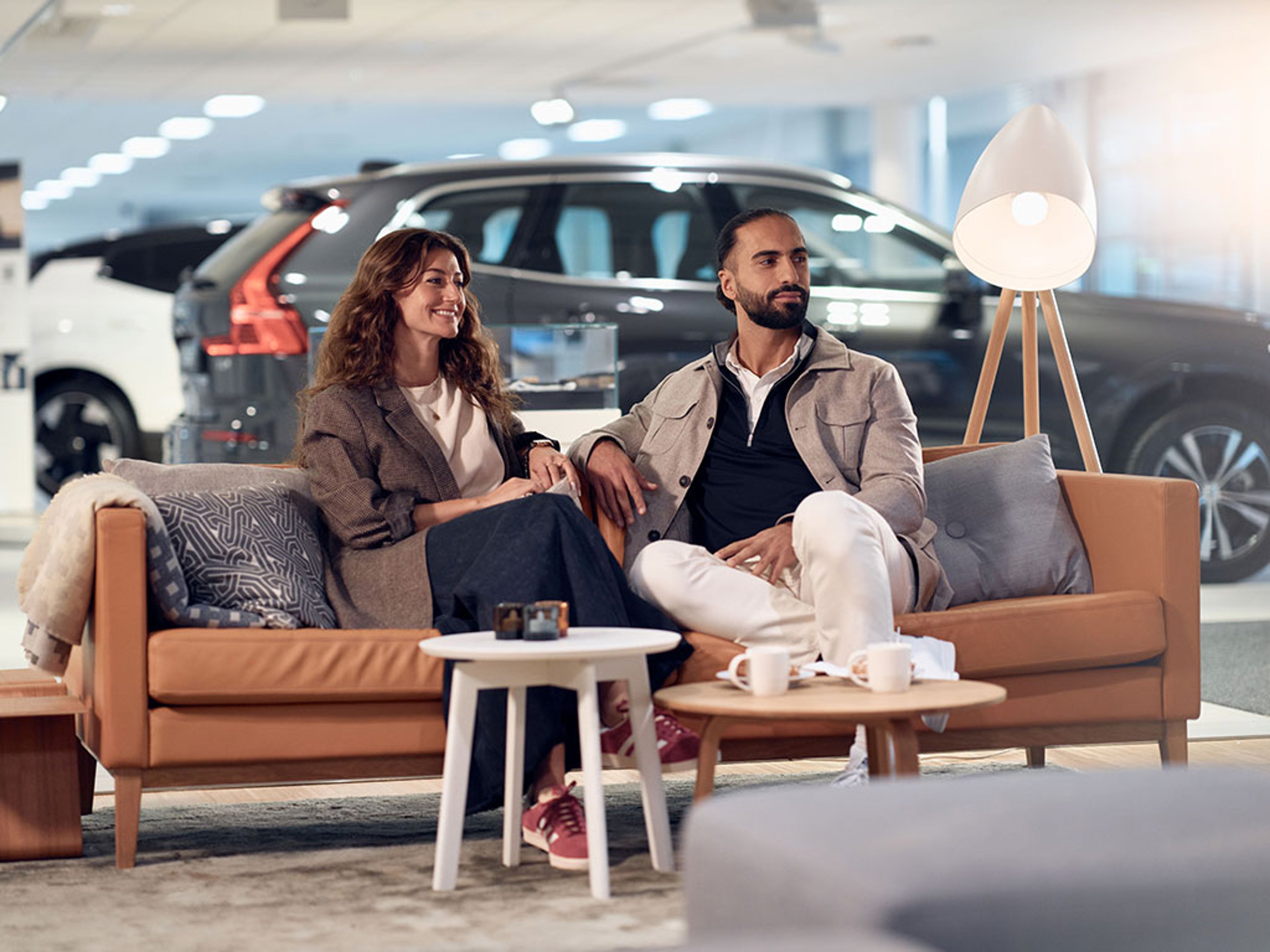 Deux personnes assises sur un canapé dans un showroom automobile avec des voitures en arrière-plan.