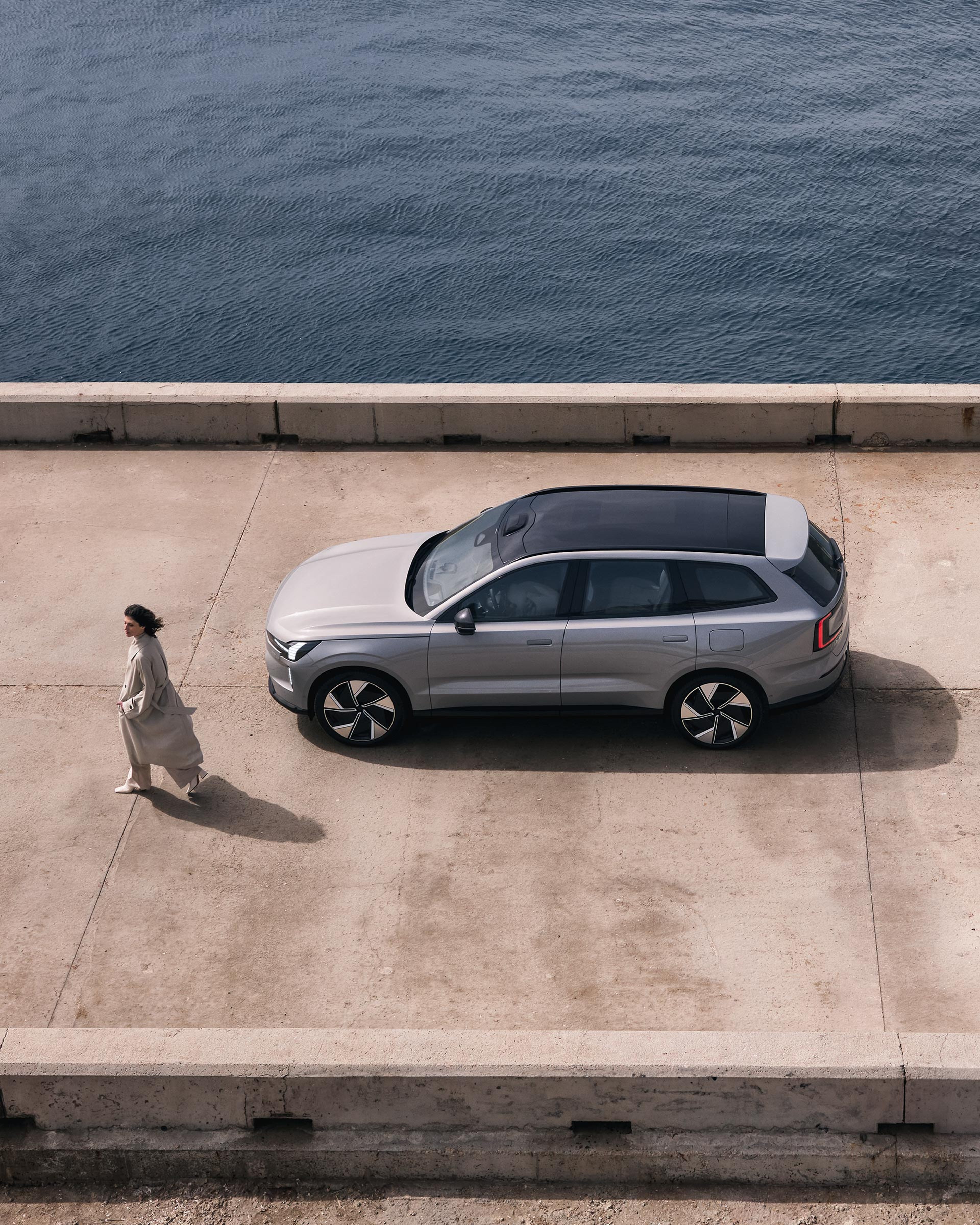 Nainen kävelee betonilaiturilla, johon on pysäköity Vapour Grey -värinen Volvo EX90 täyssähköauto.