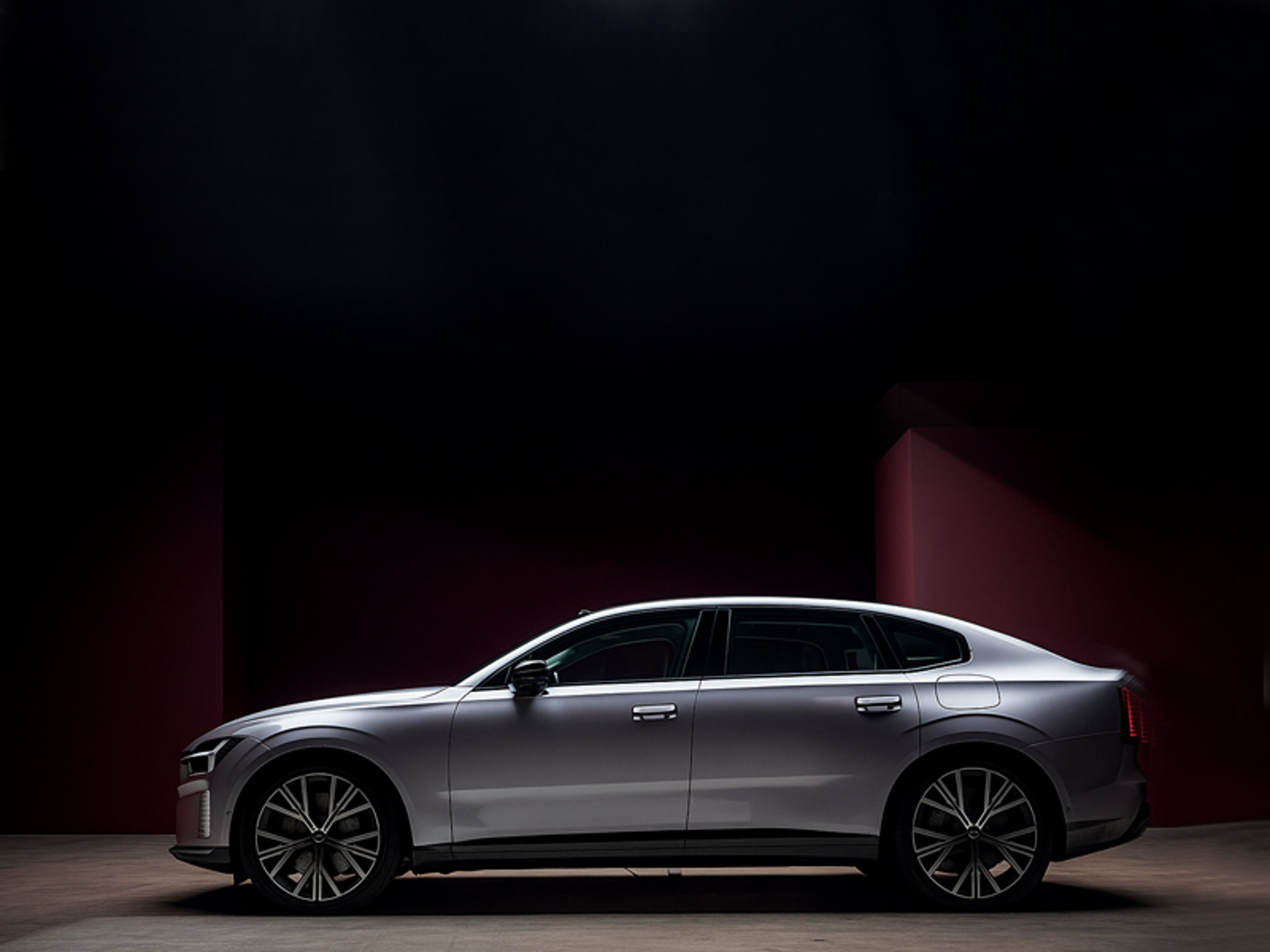 En elegant sølvfarvet Volvo bil er parkeret i et svagt oplyst miljø med en mørkerød væg. Bilen har moderne, kantede forlygter og store alufælge.