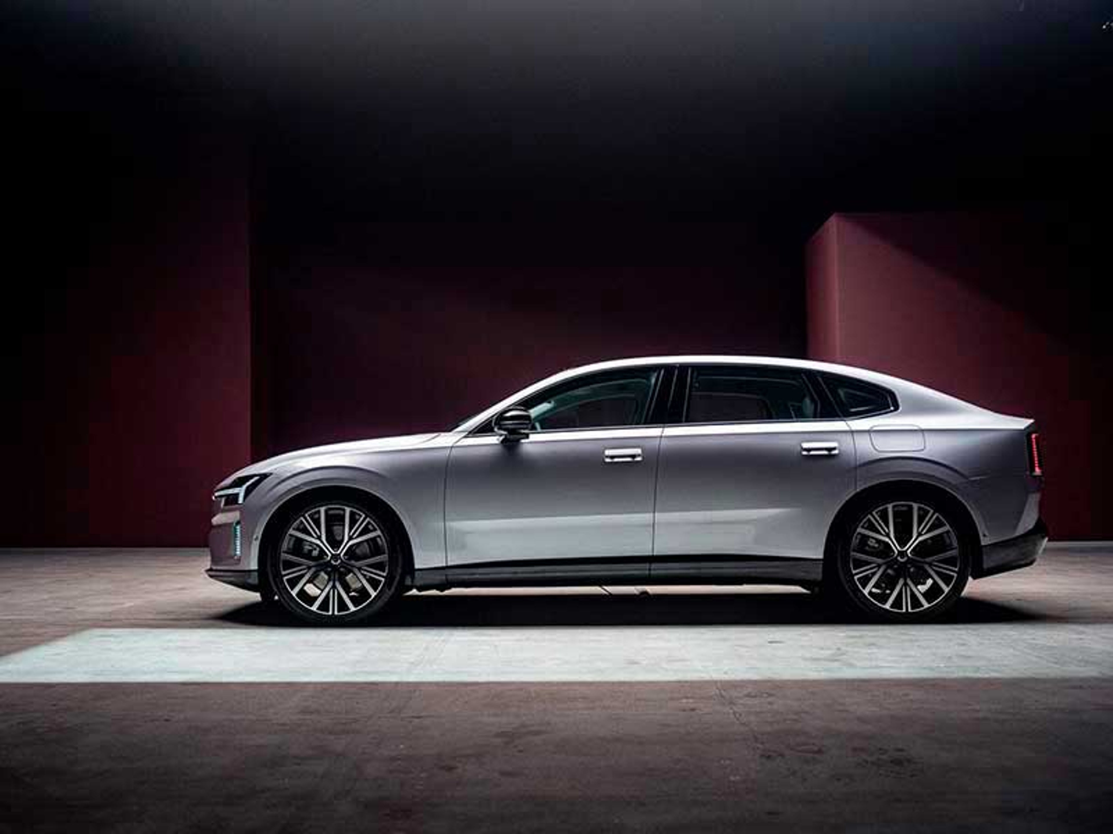 En elegant sølvfarvet Volvo bil er parkeret i et svagt oplyst miljø med en mørkerød væg. Bilen har moderne, kantede forlygter og store alufælge.