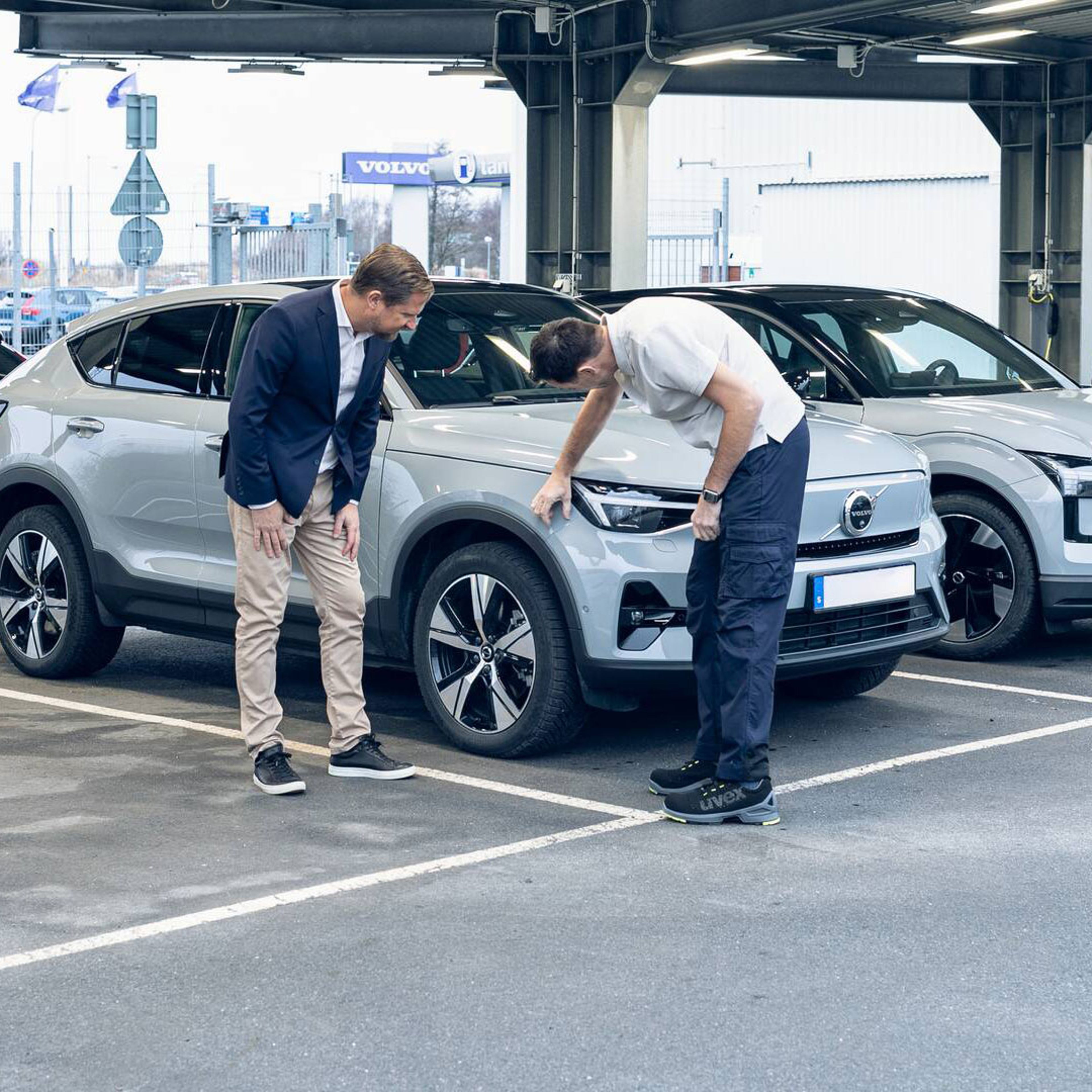 To mænd undersøger en grå Volvo bil på en parkeringsplads, under en konstruktion med industrielle elementer. Den ene peger på forhjulet, begge er fokuserede og engagerede.