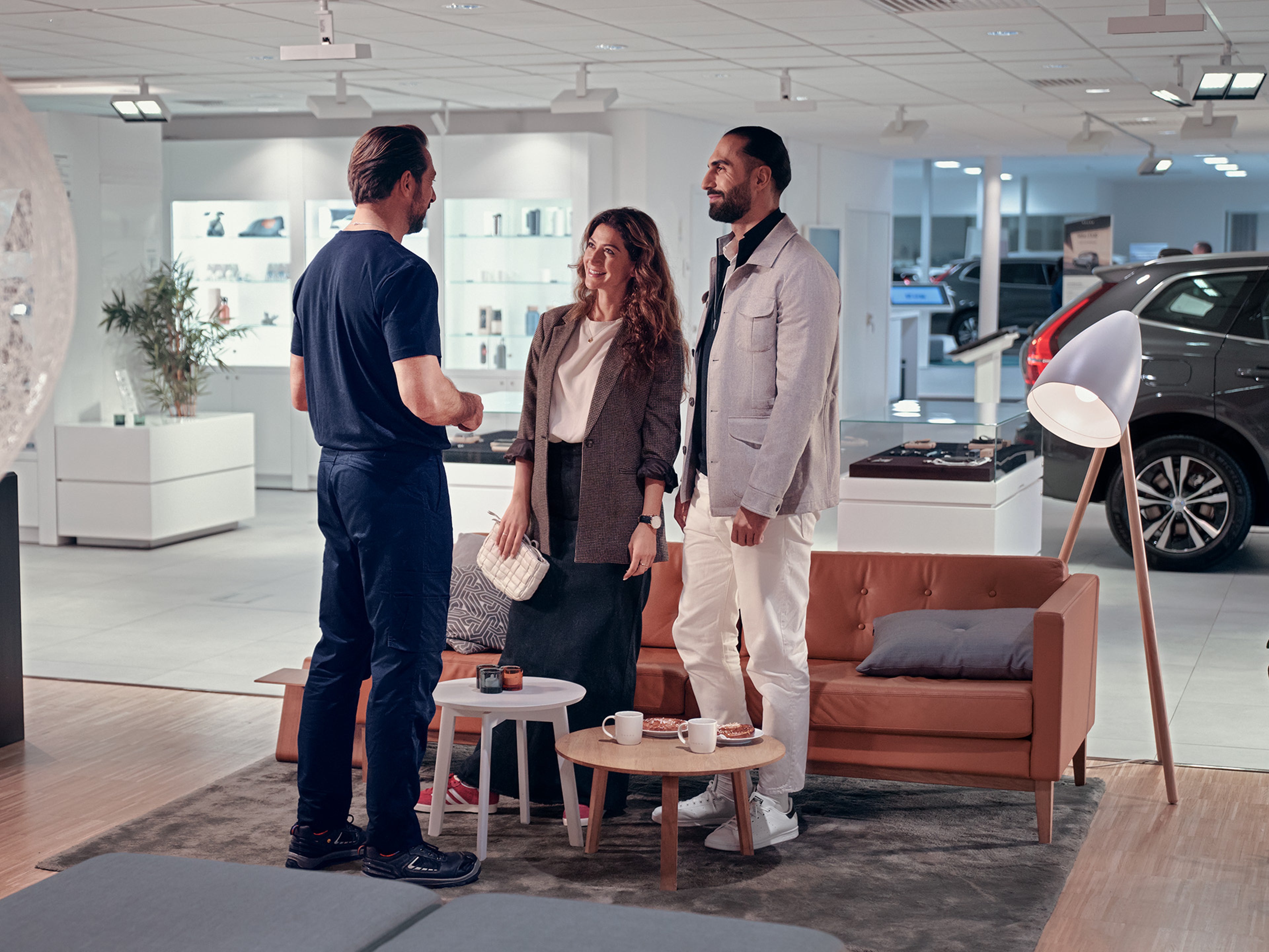 Casal, ao lado de um sofá, dentro de um concessionário, com Volvos ao fundo e um funcionário Volvo a falar com eles.