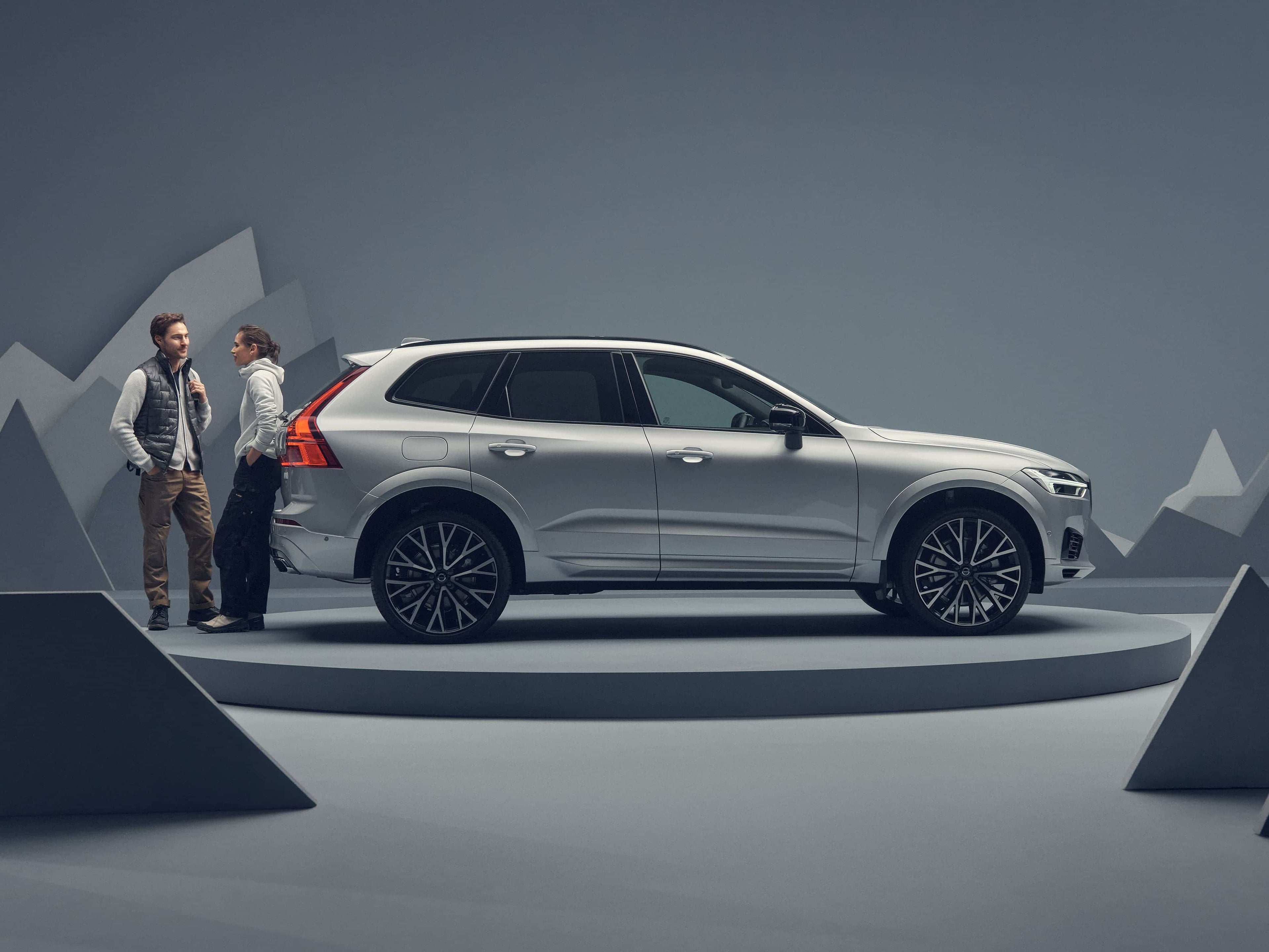 Un homme et une femme se tiennent à côté d'une Volvo XC60 argentée sur un plateau tournant dans un studio gris avec des montagnes décoratives.