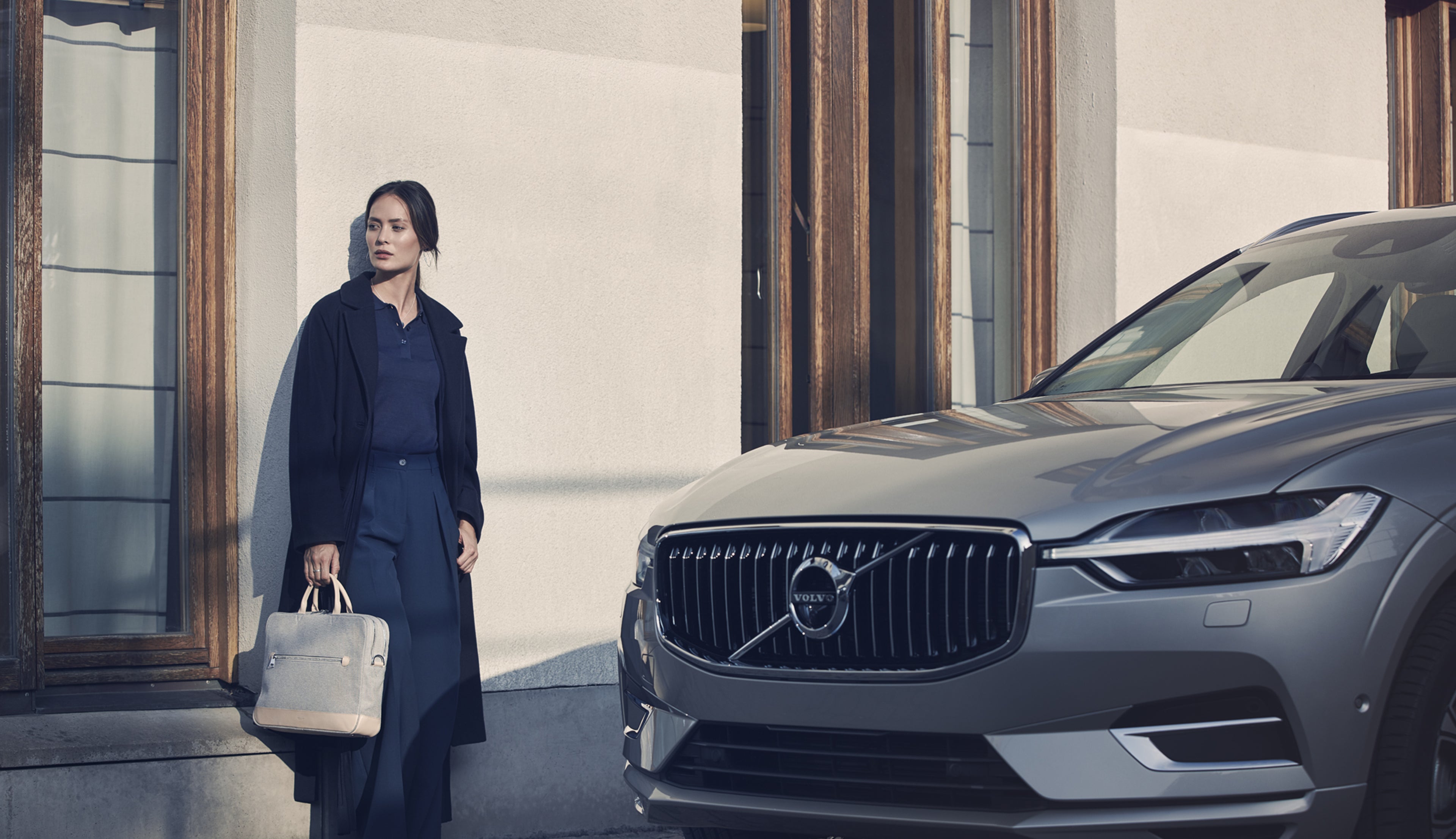 Une femme en manteau noir avec un sac beige debout à côté d'une Volvo XC60 grise devant un bâtiment à grandes fenêtres.