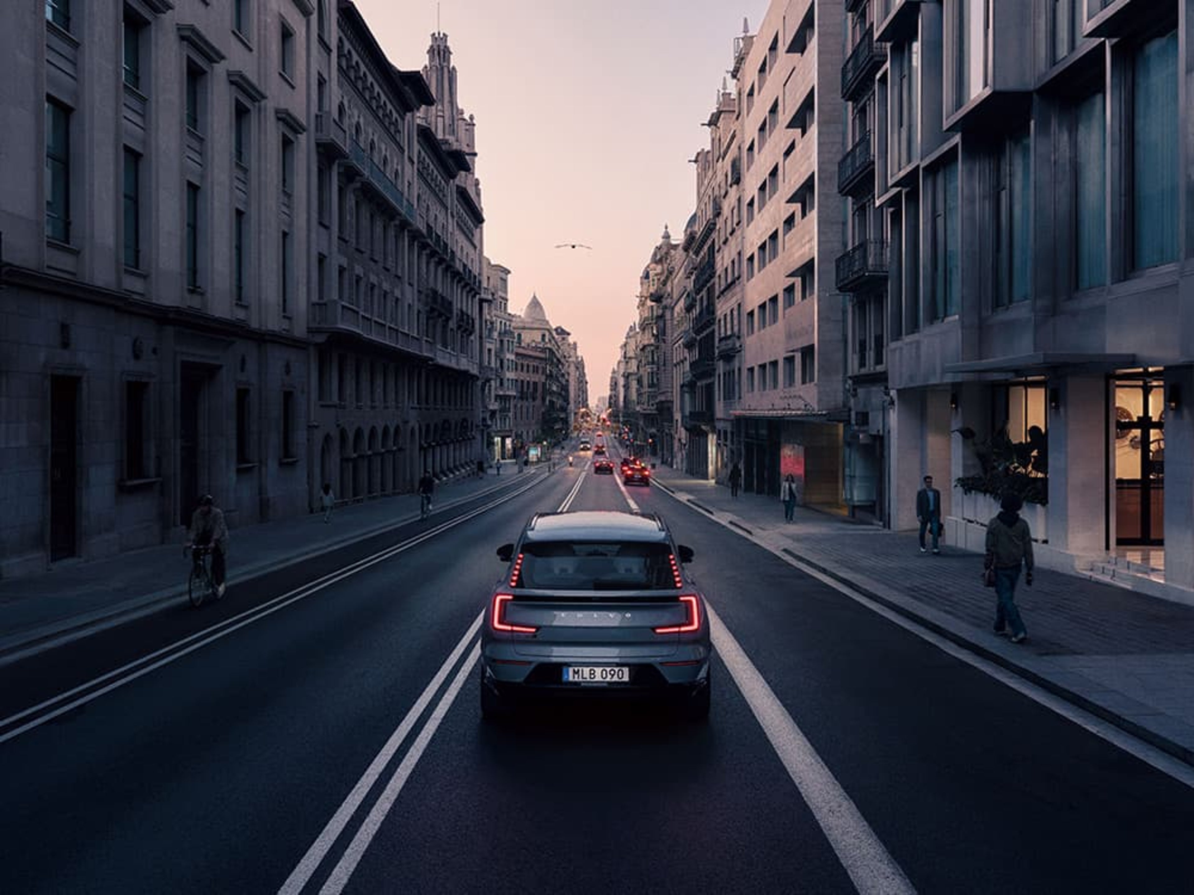 Een zilvergrijze Volvo EX90 rijdt in de schemering over een brede stadsstraat met hoge gebouwen aan weerszijden.