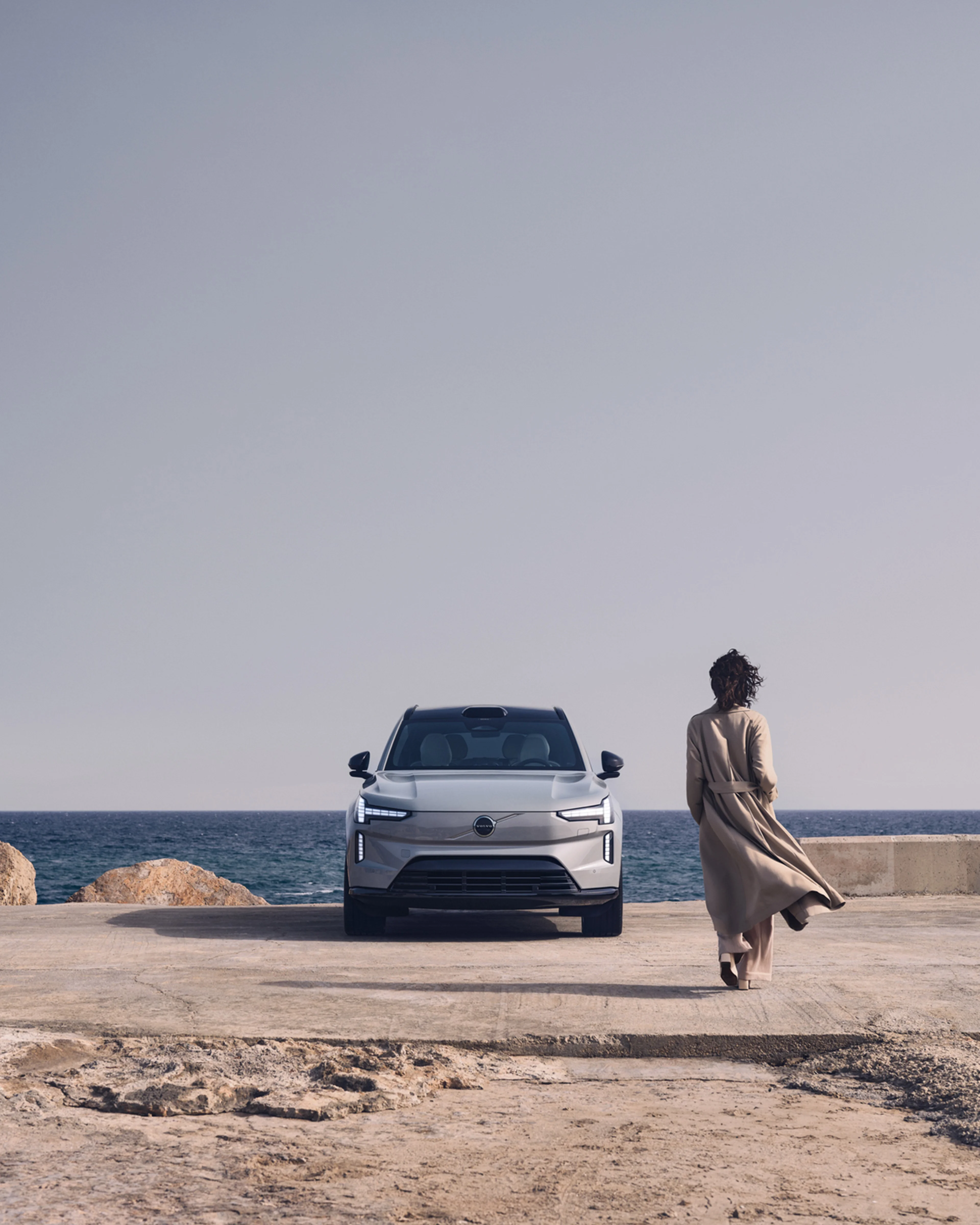 Trenssiin pukeutunut nainen kävelee kohti rantaan pysäköityä Volvo EX90 täyssähköautoa.