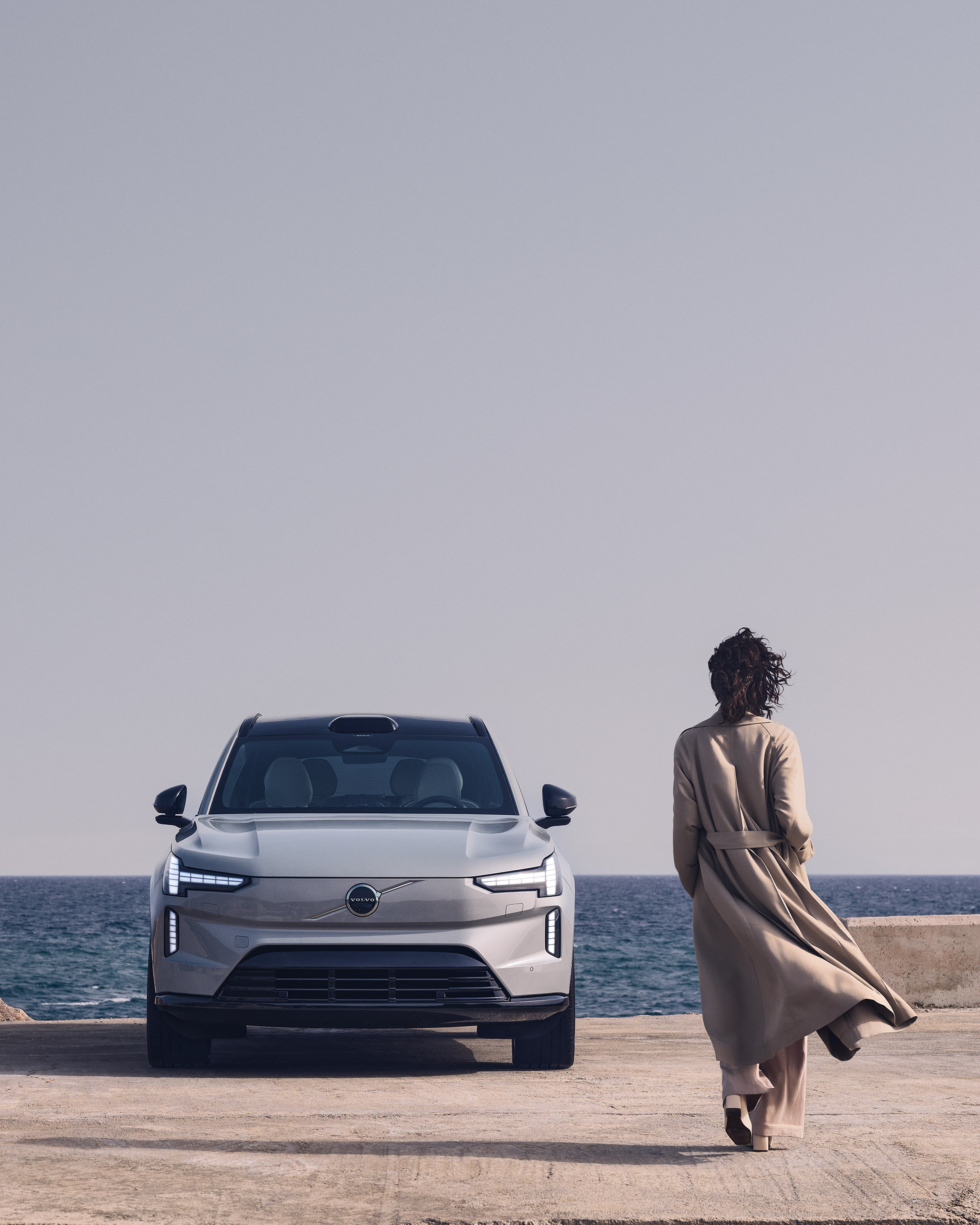 Trenssiin pukeutunut nainen kävelee kohti rantaan pysäköityä Volvo EX90 täyssähköautoa.
