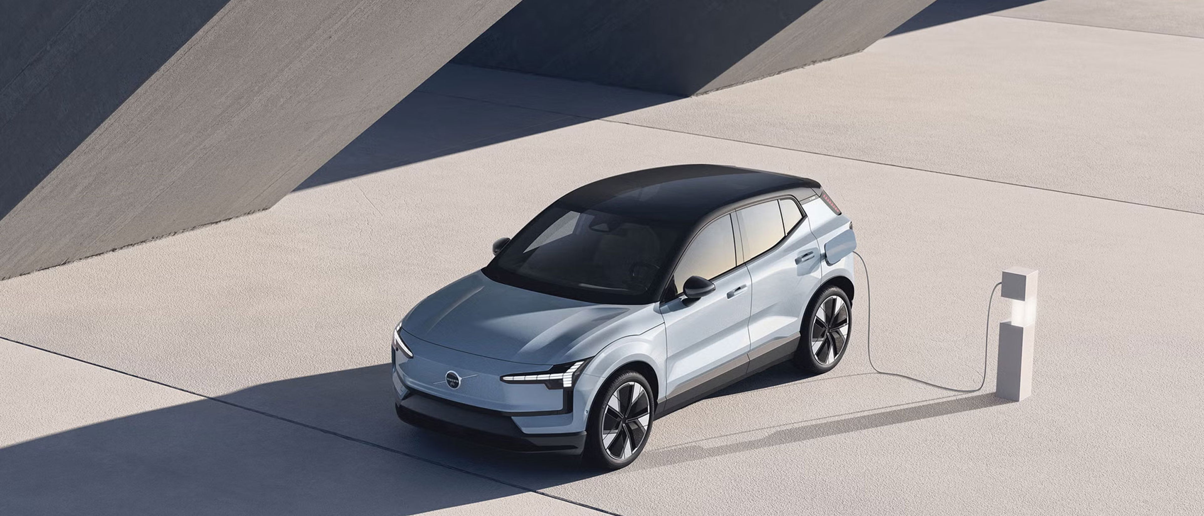 Gornji pogled na luksuzni elektrificirani Volvo XC90 SUV u boji Bright Dusk, parkiran na modernoj betonskoj površini ispred zida s rebrastom teksturom, naglašavajući elegantan dizajn i ekološku orijentaciju.