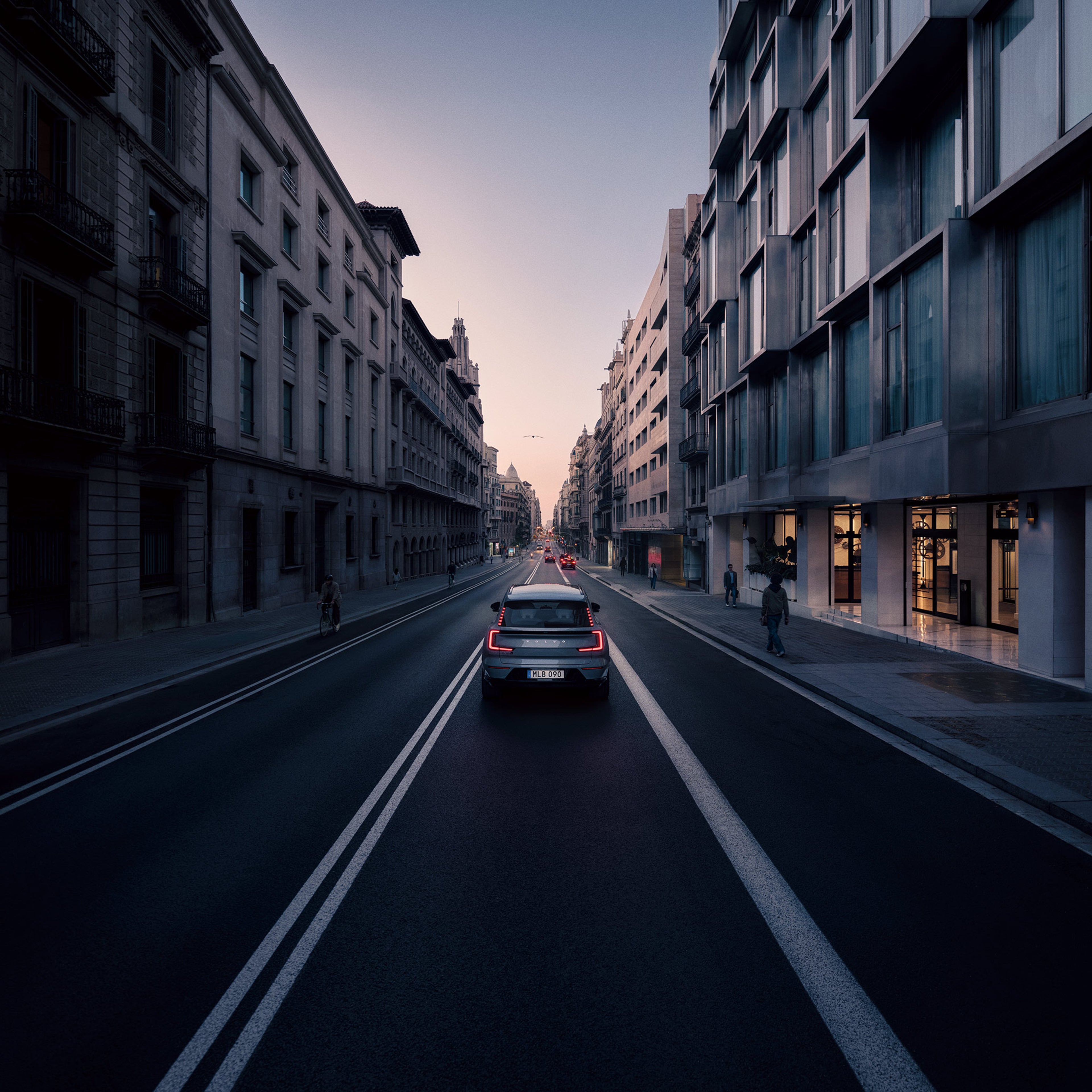 Takaapäin kuvattu Vapour grey -värinen Volvo EX90 täyssähköinen katumaasturi ajaa kaupungissa kadulla kohti horisonttia iltahämärässä.