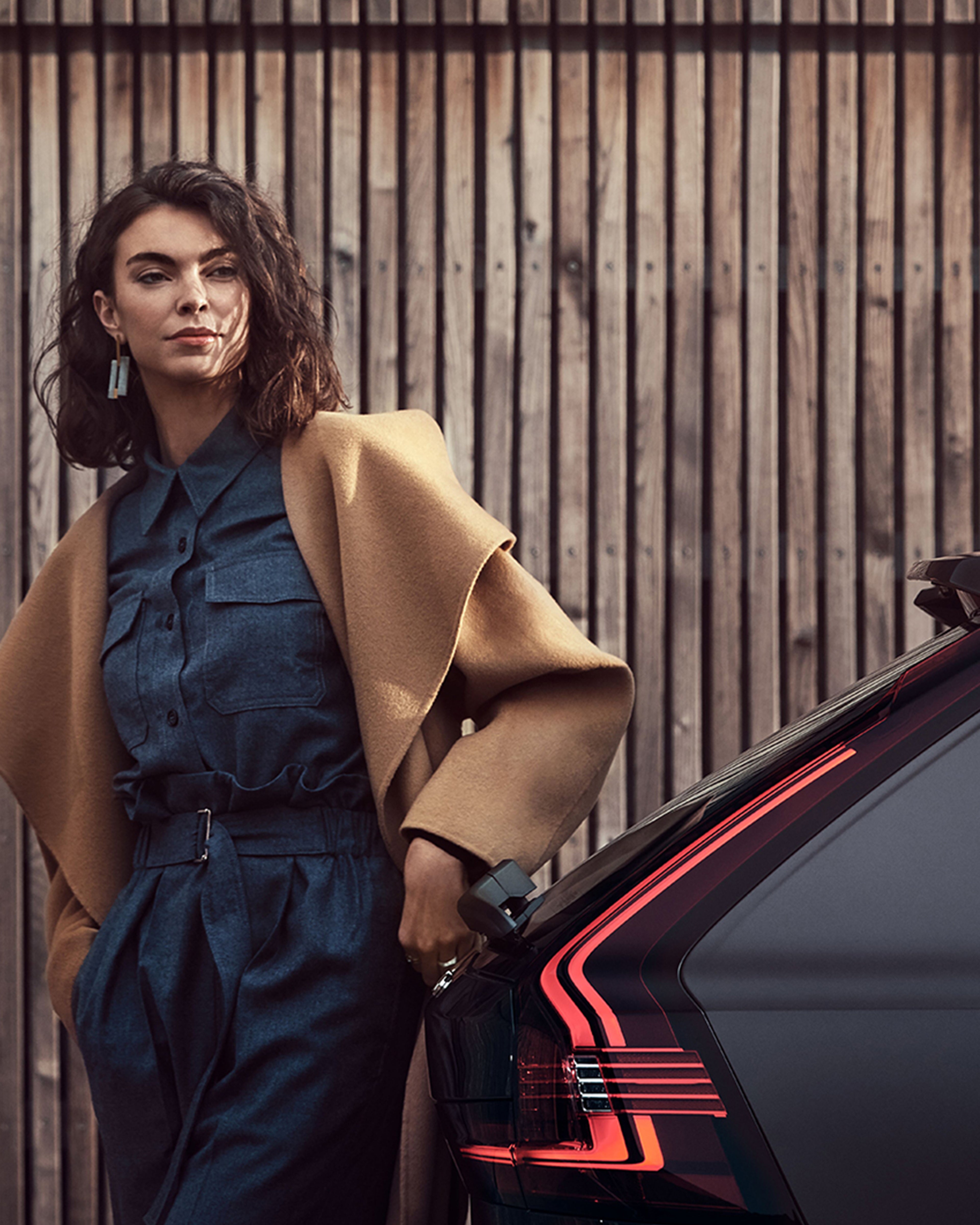 Eine junge Frau in einem braunen Mantel steht am Heck eines Volvo