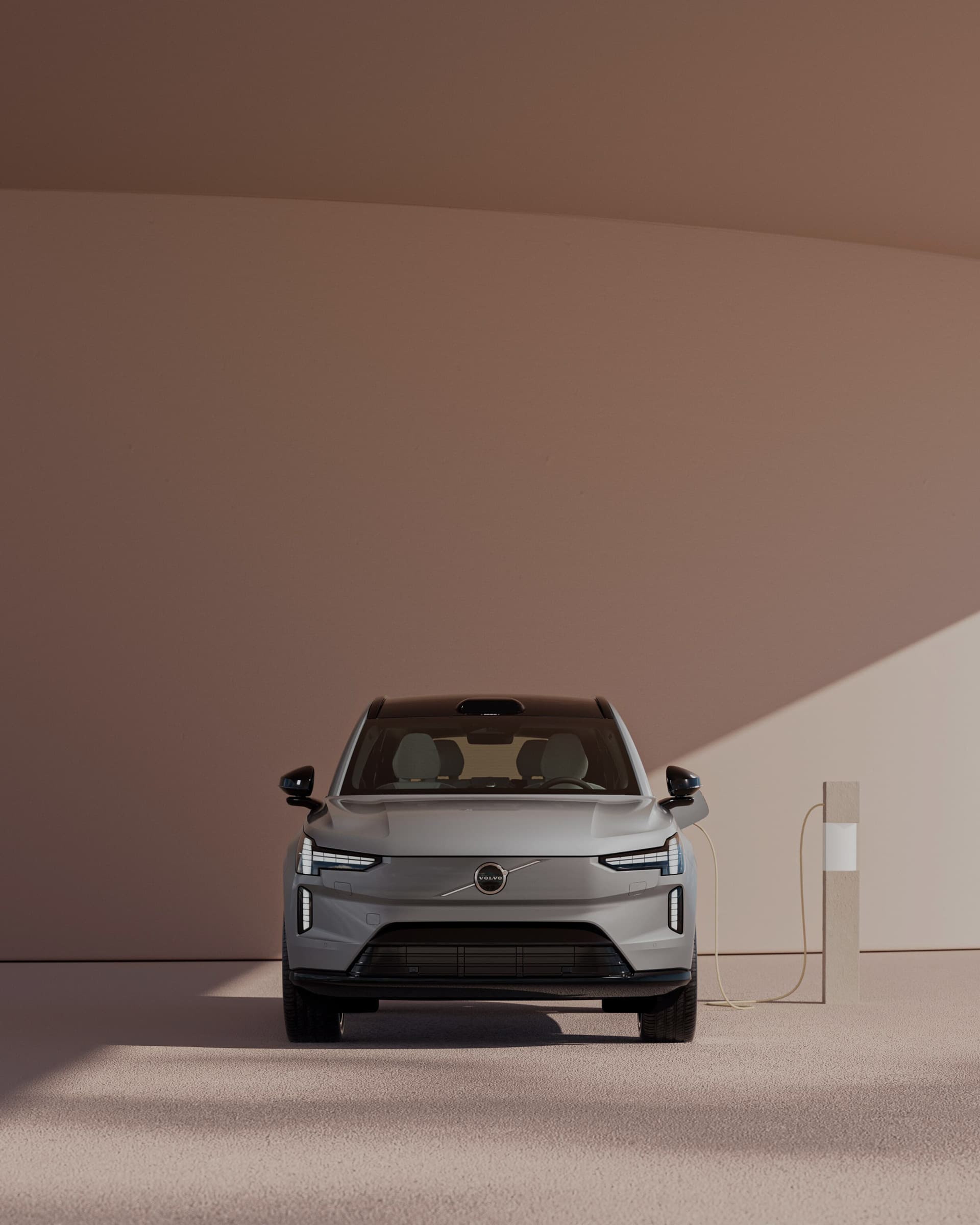 Avant d'un Volvo EX90 gris connecté à une borne de recharge sur fond orange pastel.
