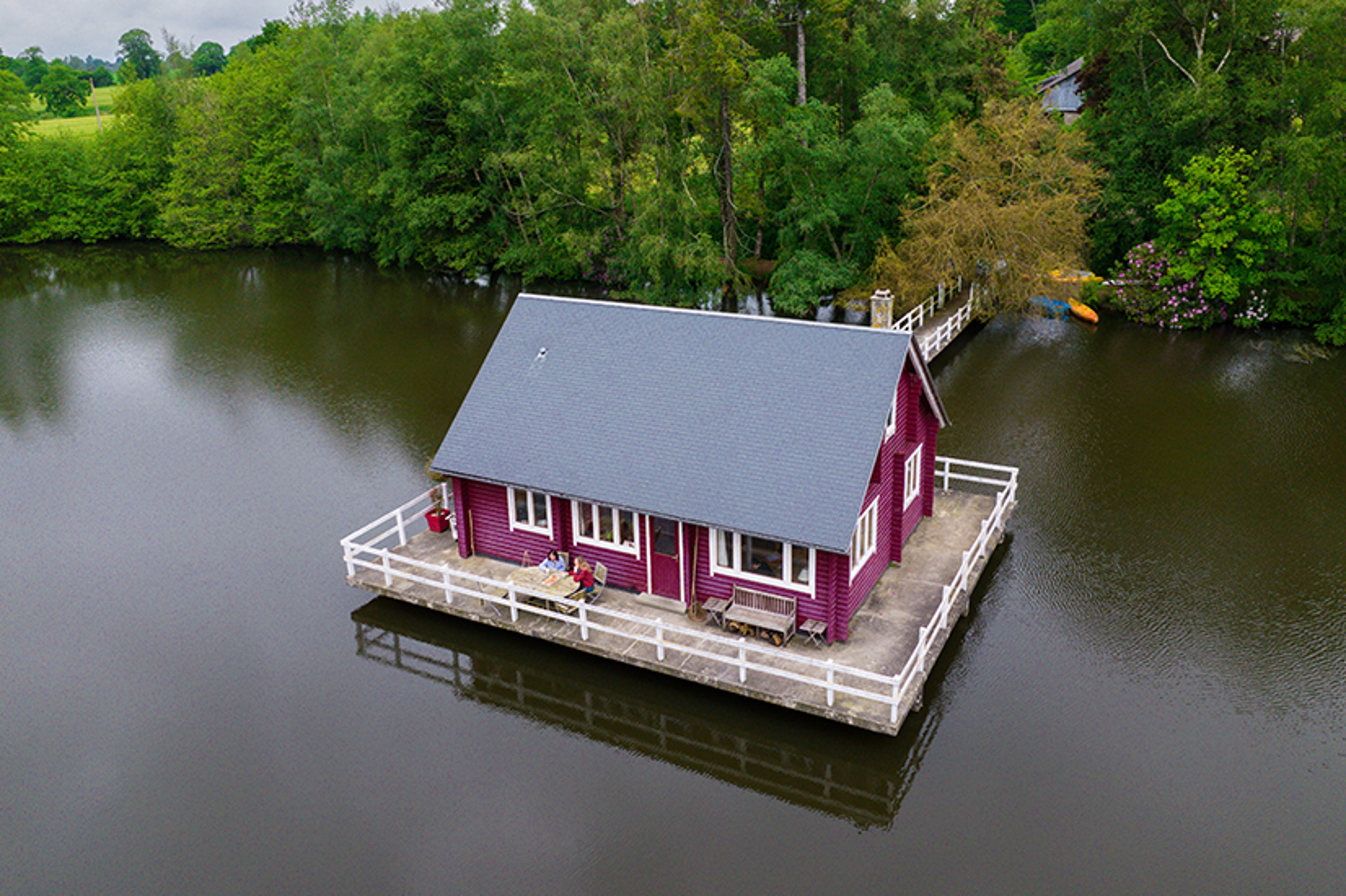 Maison rouge au bord d’un lac forestier, vue aérienne avec terrasse et mobilier extérieur.