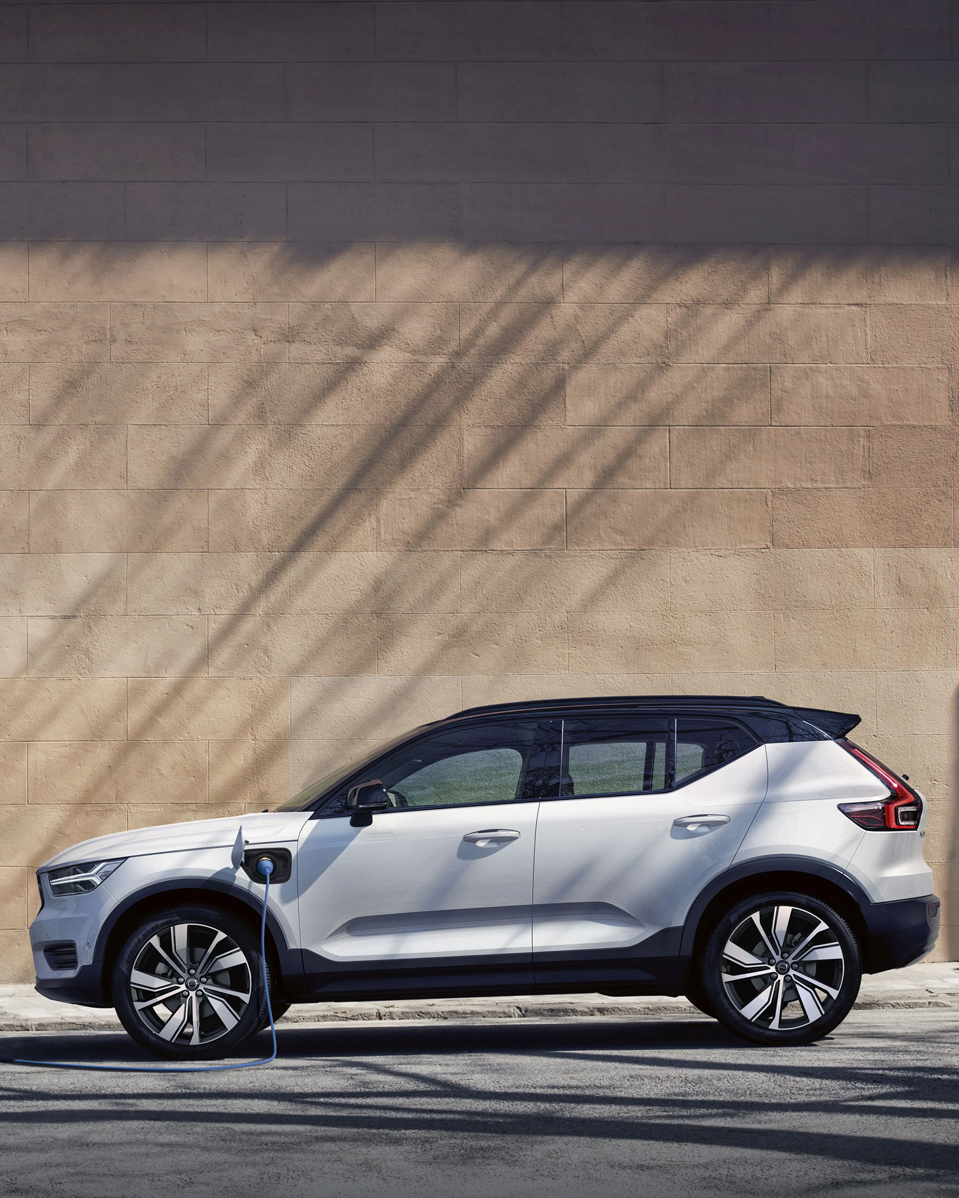 Voiture électrique blanche en charge devant un mur beige, ombres d’arbres visibles.