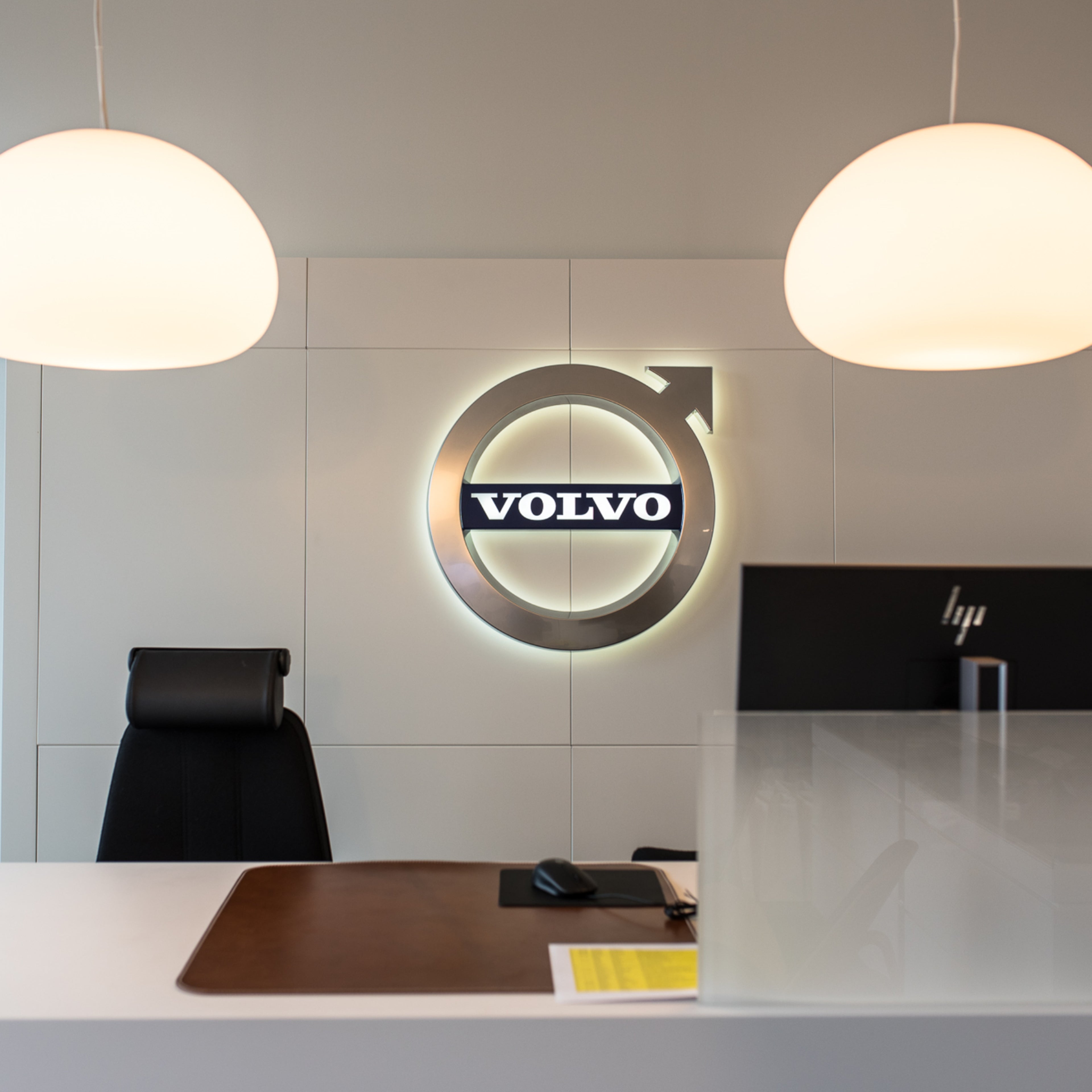Ein großes Volvo Emblem hängt in einem Büro, zu sehen sind der Schreibtisch mit einer Mausunterlage und der Maus, der Schreibtischstuhl und ein HP Bildschirm.
