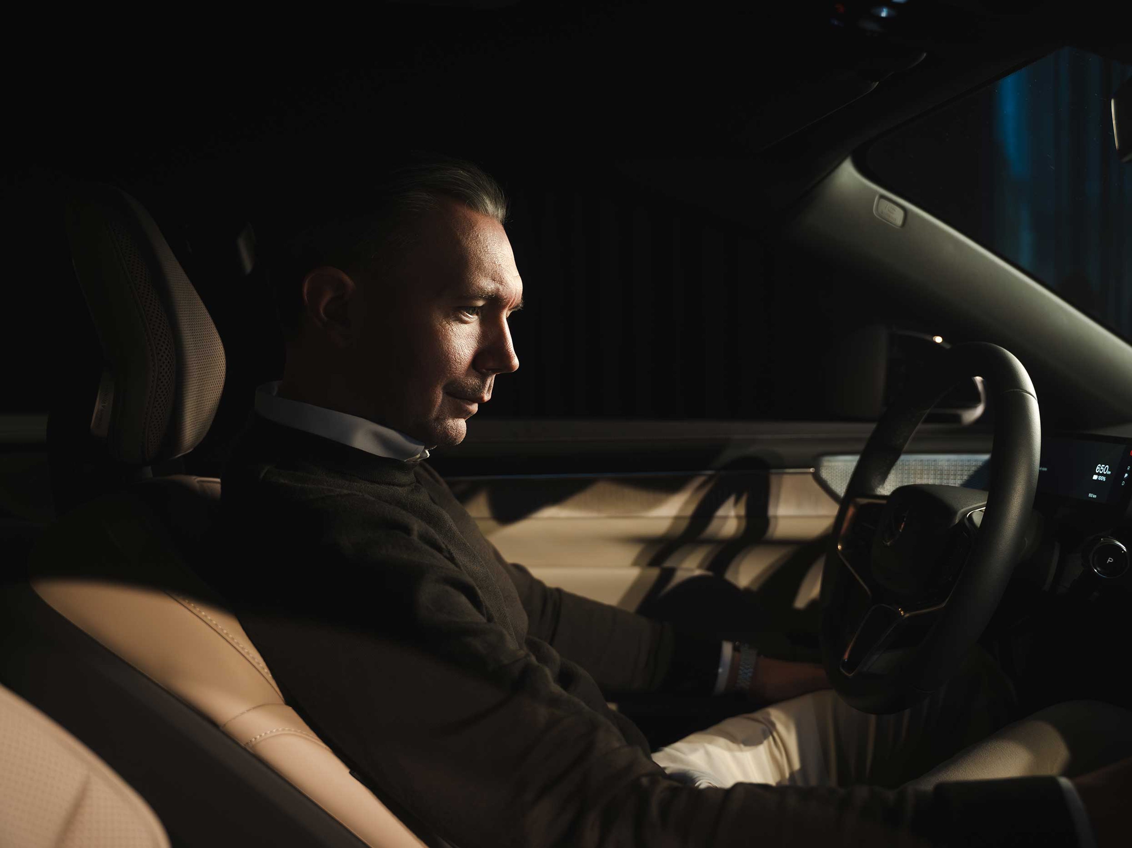 En mand i et svagt oplyst bilinteriør, iført en mørk sweater, kigger eftertænksomt ned. Bløde skygger og varm belysning skaber en kontemplativ stemning.