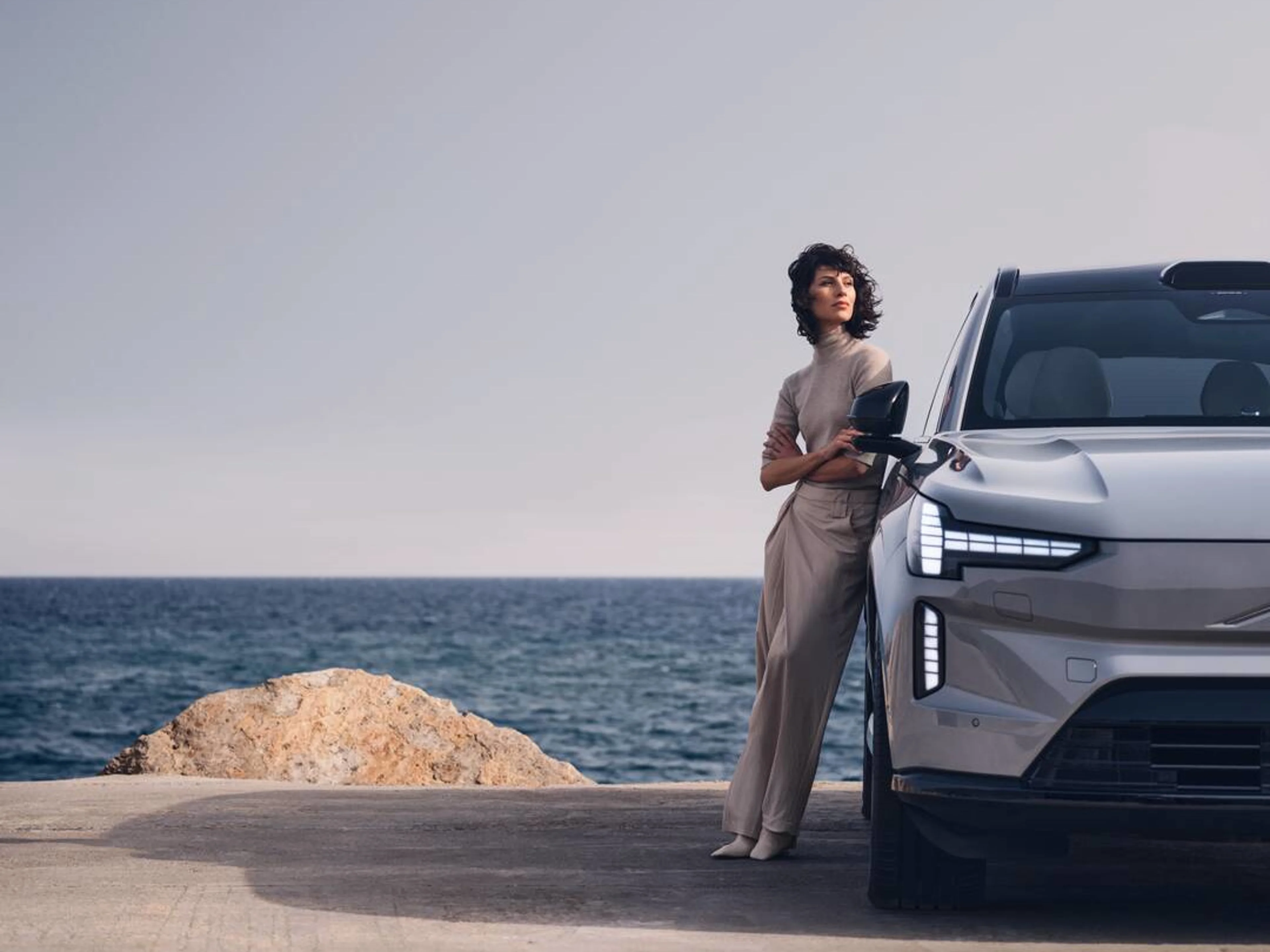 A stylish person leans against a Volvo EX90, overlooking a serene ocean landscape under a clear sky.