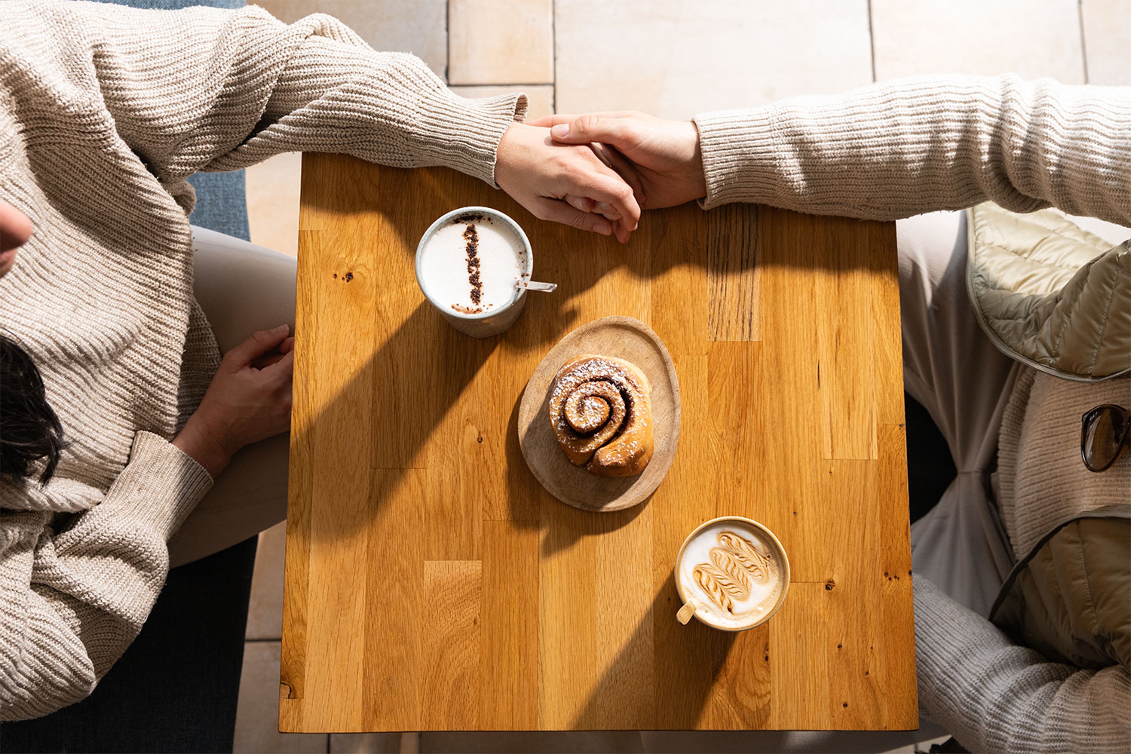 Deux personnes se tiennent la main autour d’une table en bois avec cafés et roulé à la cannelle.
