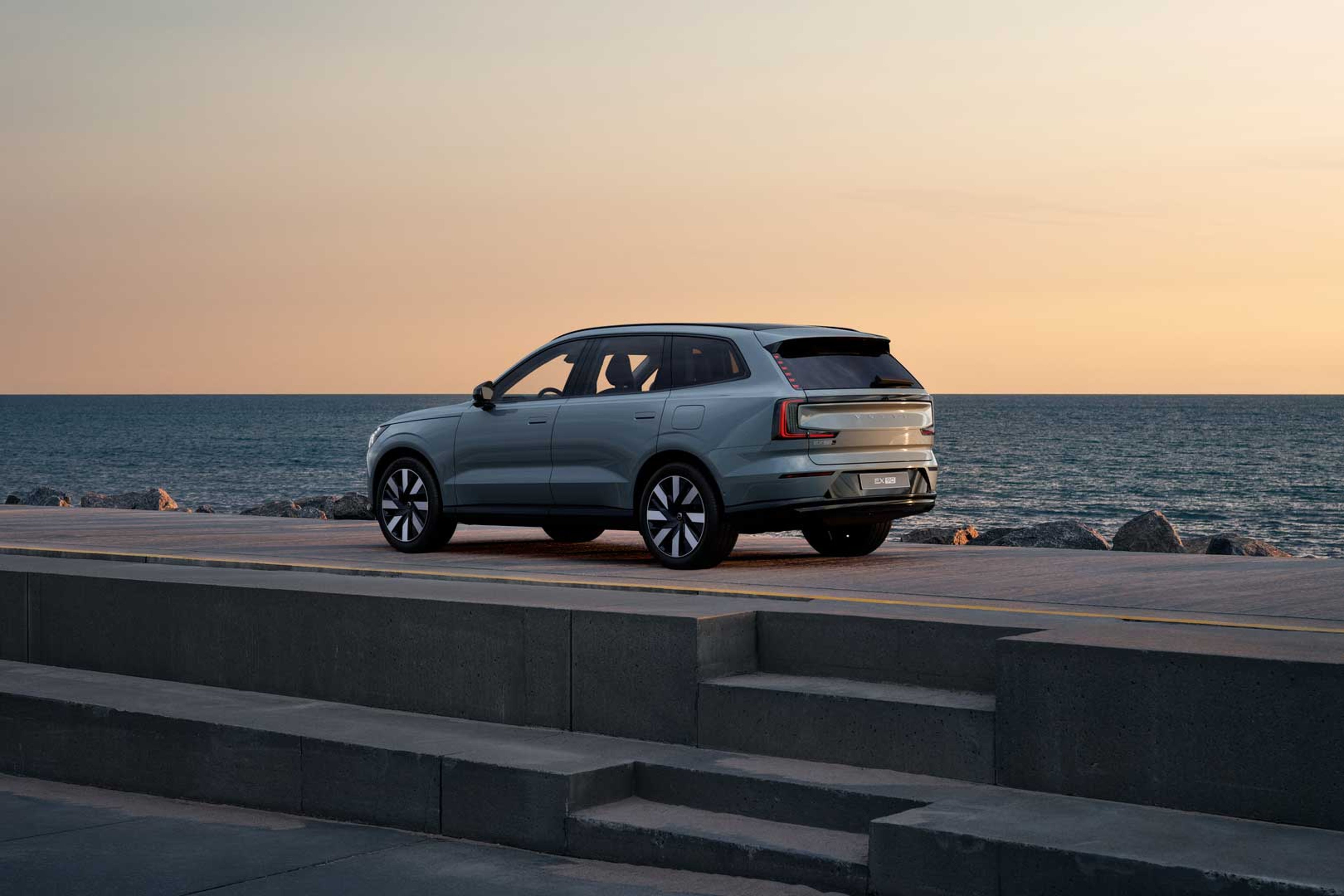 Elegant grå Volvo EX90 SUV parkeret ved havet ved solnedgang på en cementplatform. Himlen har en blød farvegradient af varme nuancer, som skaber en rolig og fredfyldt stemning.