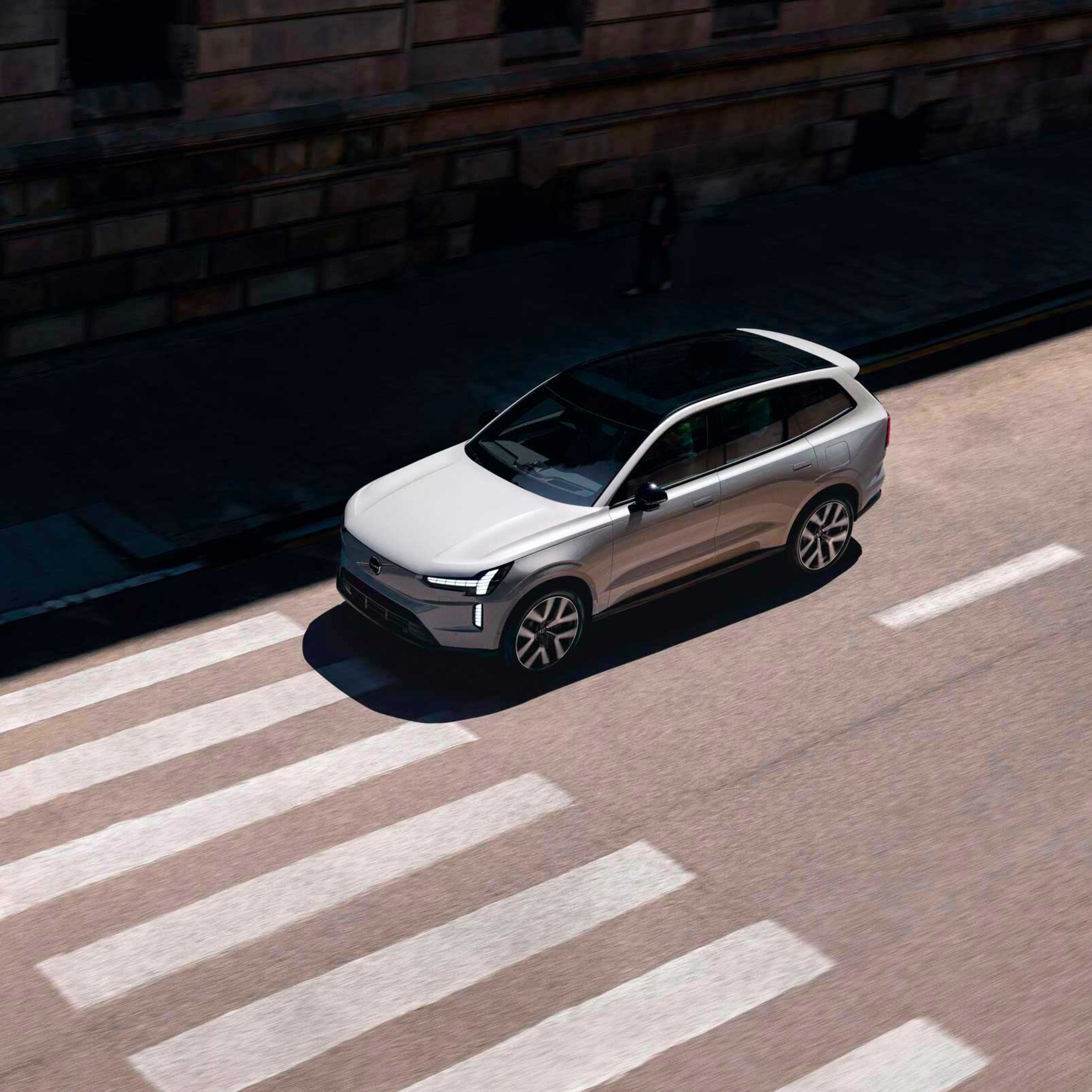 En sølvfarvet Volvo SUV kører over et fodgængerfelt på en solrig dag. Køretøjets elegante design står i kontrast til den skyggefulde, urbane baggrund og skaber en dynamisk scene.