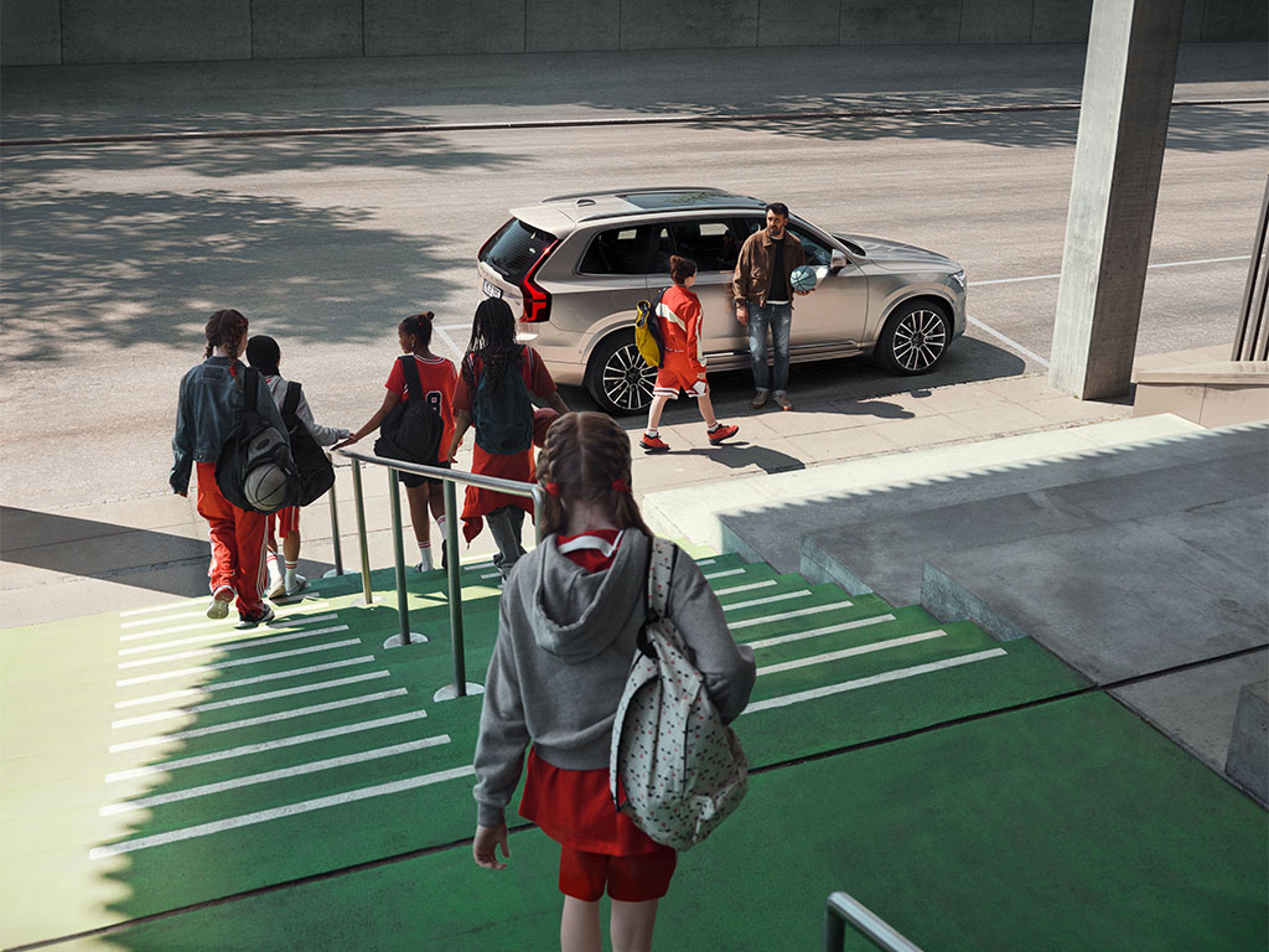Un homme avec un ballon de basket, adossé à son XC90 gris clair, attend des jeunes qui sortent du gymnase