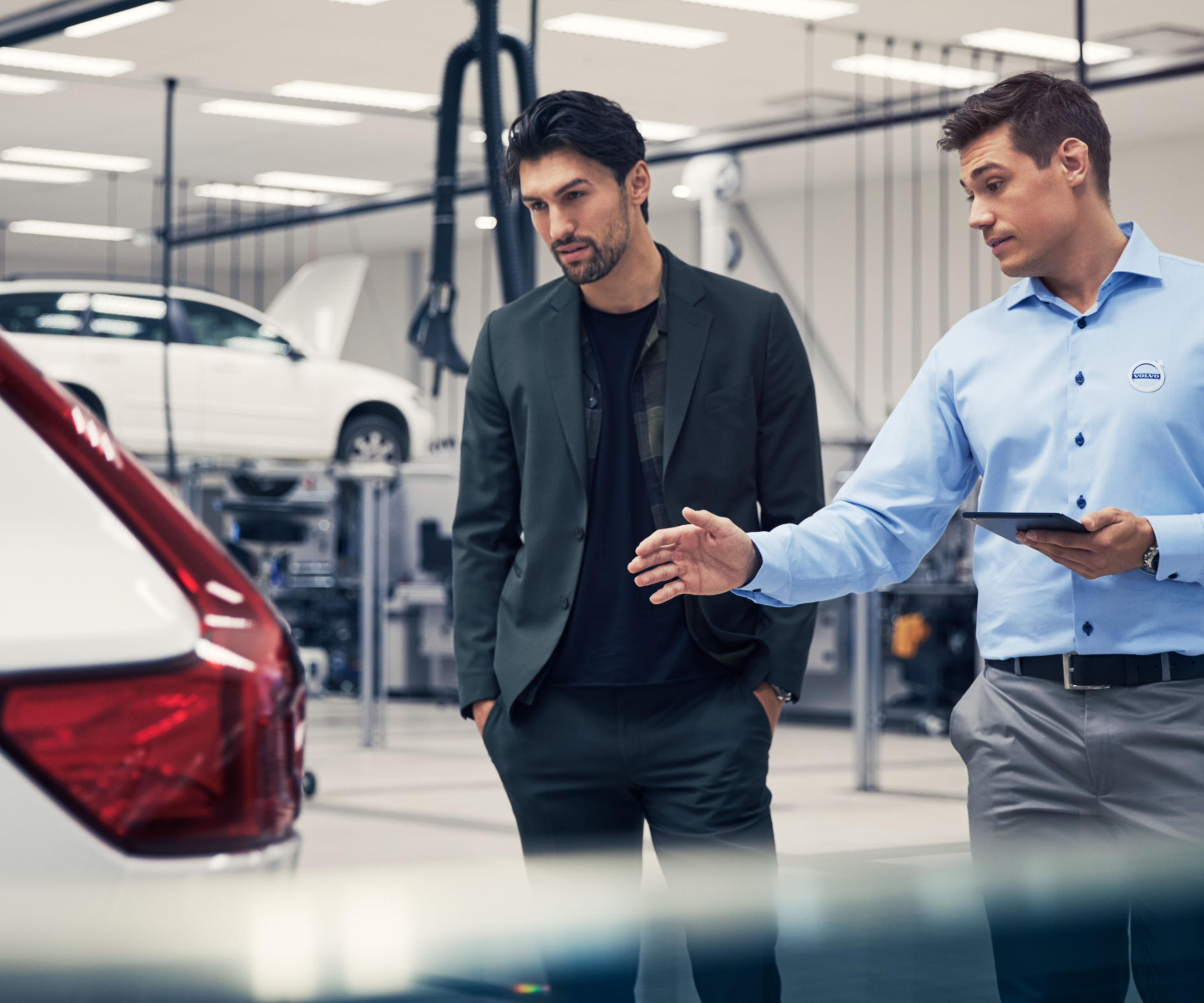 Zwei Männer befinden sich in einer Werkstatt und unterhalten sich über ein Fahrzeug.