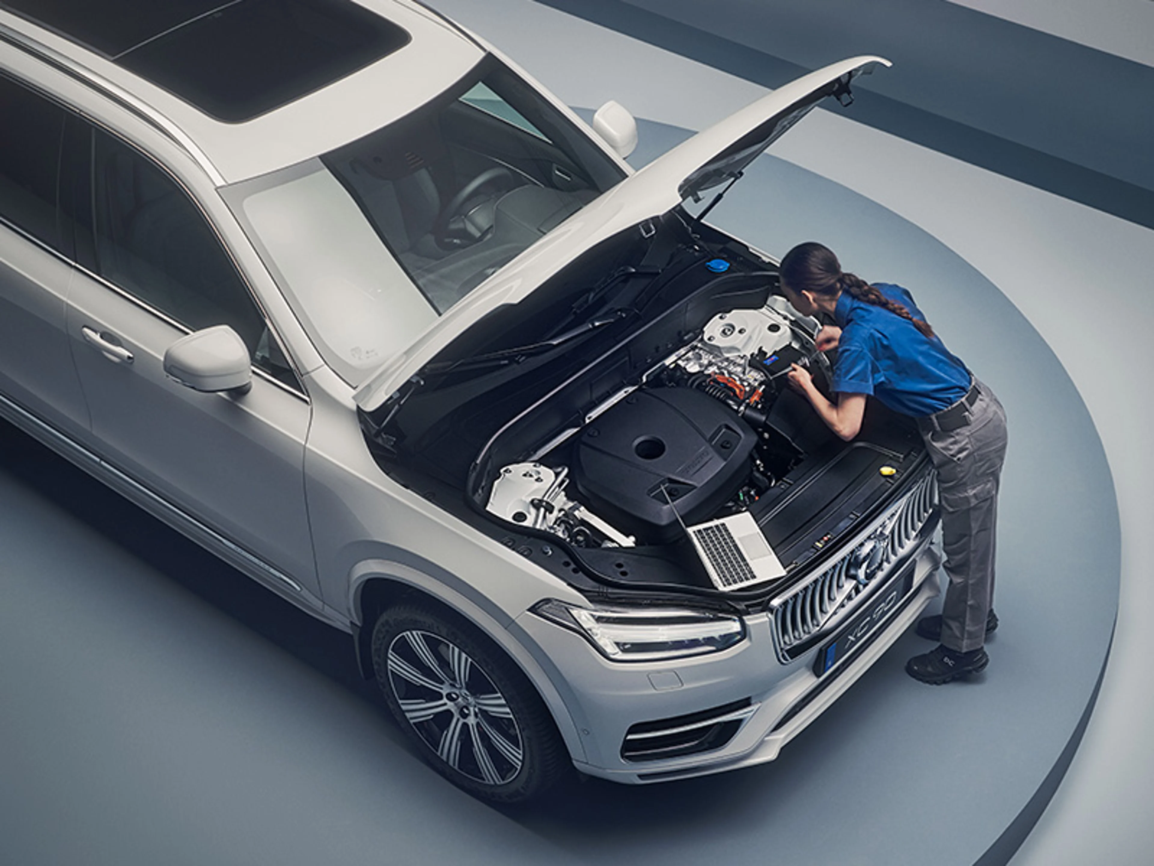Une Volvo XC90 blanche avec le capot ouvert, une mécanicienne se tient devant la voiture et travaille sur le moteur avec un ordinateur portable à côté.