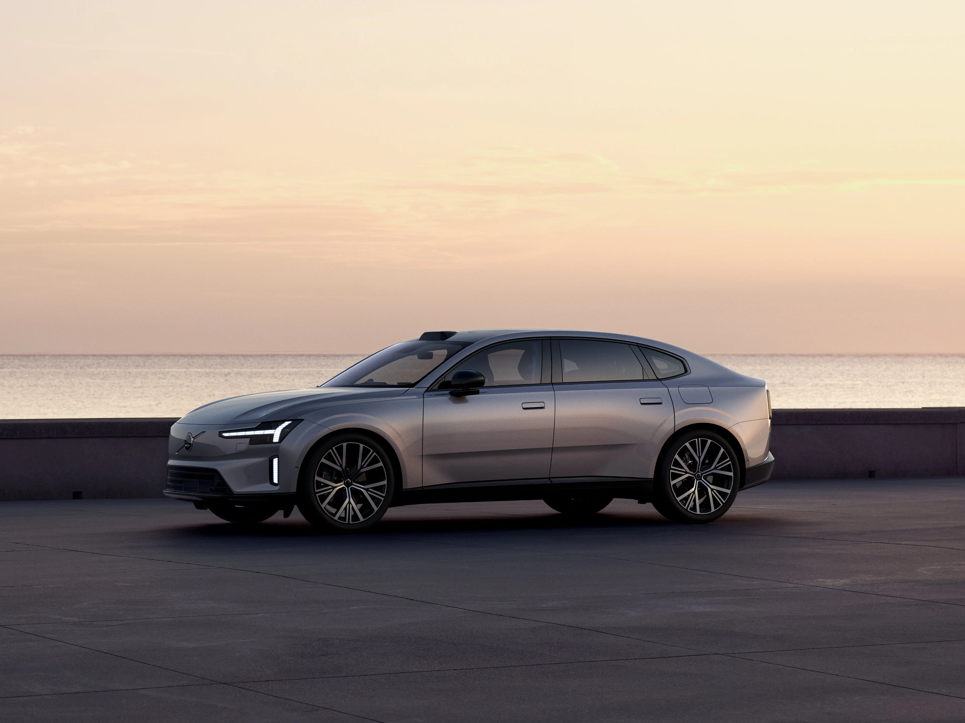 Ein silberner Volvo ES90 parkt bei Zwielicht an einem Küstenstreifen vor dem Meer.